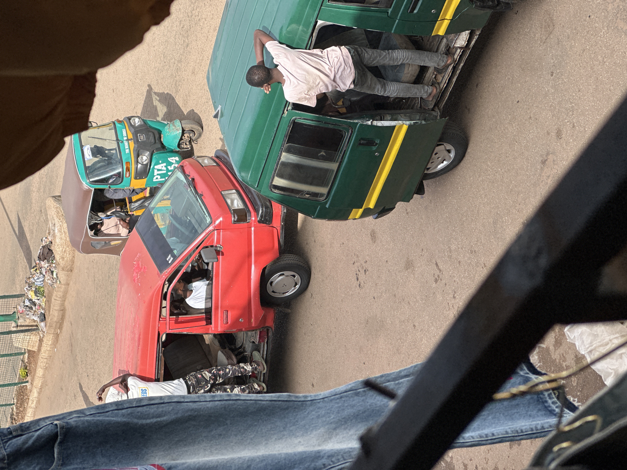 In the city of Bukuru, Nigeria, a street scene unfolds with multiple vehicles and pedestrians. A red van is parked, its driver's side door open, revealing a person in the driver's seat. A man in a white t-shirt and patterned pants stands near the open door, his arm raised. To the right, a green van has its passenger door open, with a person standing on the step. A person is leaning on the roof of the green van. In the background, a tri-scooter, also known as an auto-rickshaw, is visible, with a person inside. The street surface is paved, and there is a curb with scattered trash along it. A portion of blue denim jeans hangs into the foreground on the left side of the frame, partially obscuring the view.