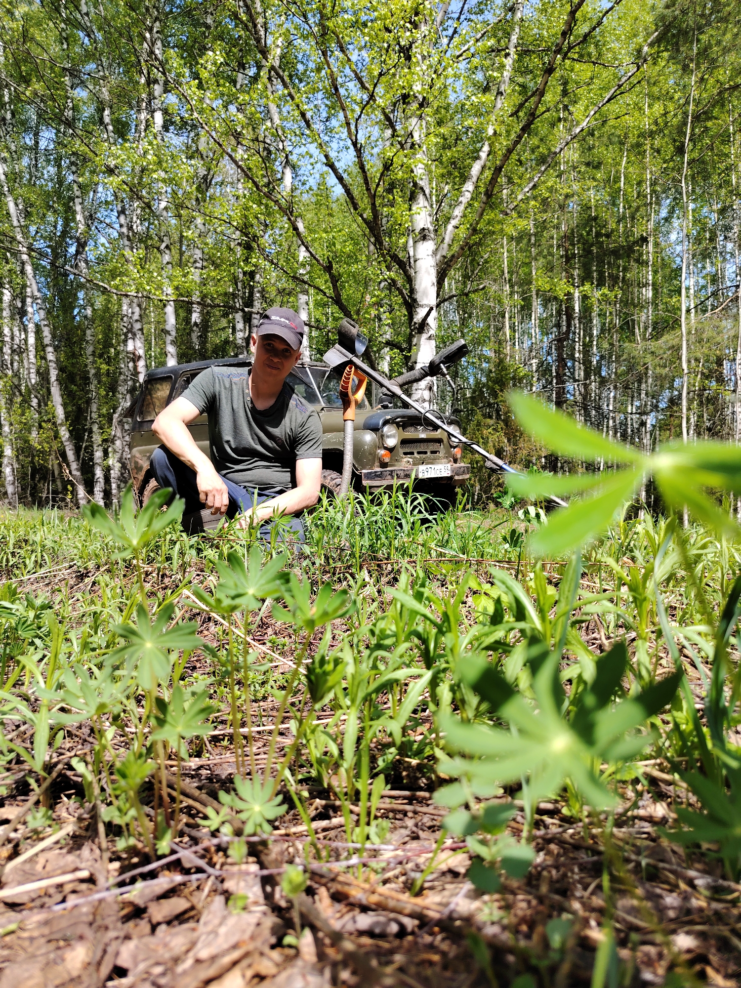 The image shows a man kneeling in a grassy area with trees in the background. Behind him is an olive green SUV with the license plate "P 971 CE 55". A metal detector and a shovel are resting against the front of the SUV. The location is Markovskii, Russia, as indicated by the license plate. The ground is covered with loose soil and small green plants. Some larger green leafy plants are in the foreground. The man is wearing a dark T-shirt, jeans, and a baseball cap. The trees in the background are mostly birch trees with some evergreen trees interspersed. The sky is visible through the trees, appearing blue.