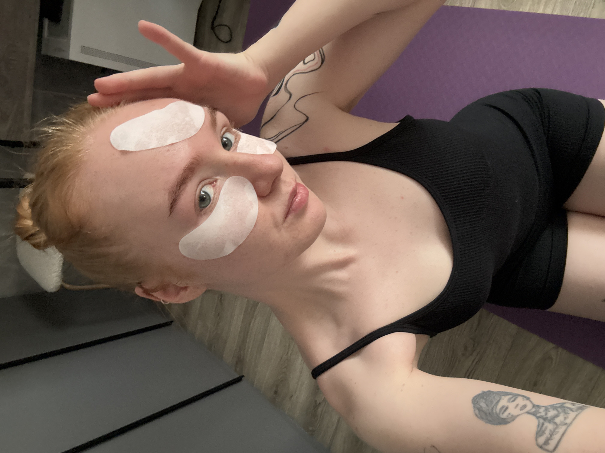 The image shows a young woman with red hair pulled up in a bun, wearing a black tank top and shorts. She has several white under-eye patches and one on her forehead. She is looking at the camera with her right hand raised to her forehead. She has tattoos on her left arm and shoulder. She is on a purple yoga mat, with a wooden floor visible in the background. The scene is indoors, likely a personal space. The location is not identifiable as Chaikovsky, Russia.