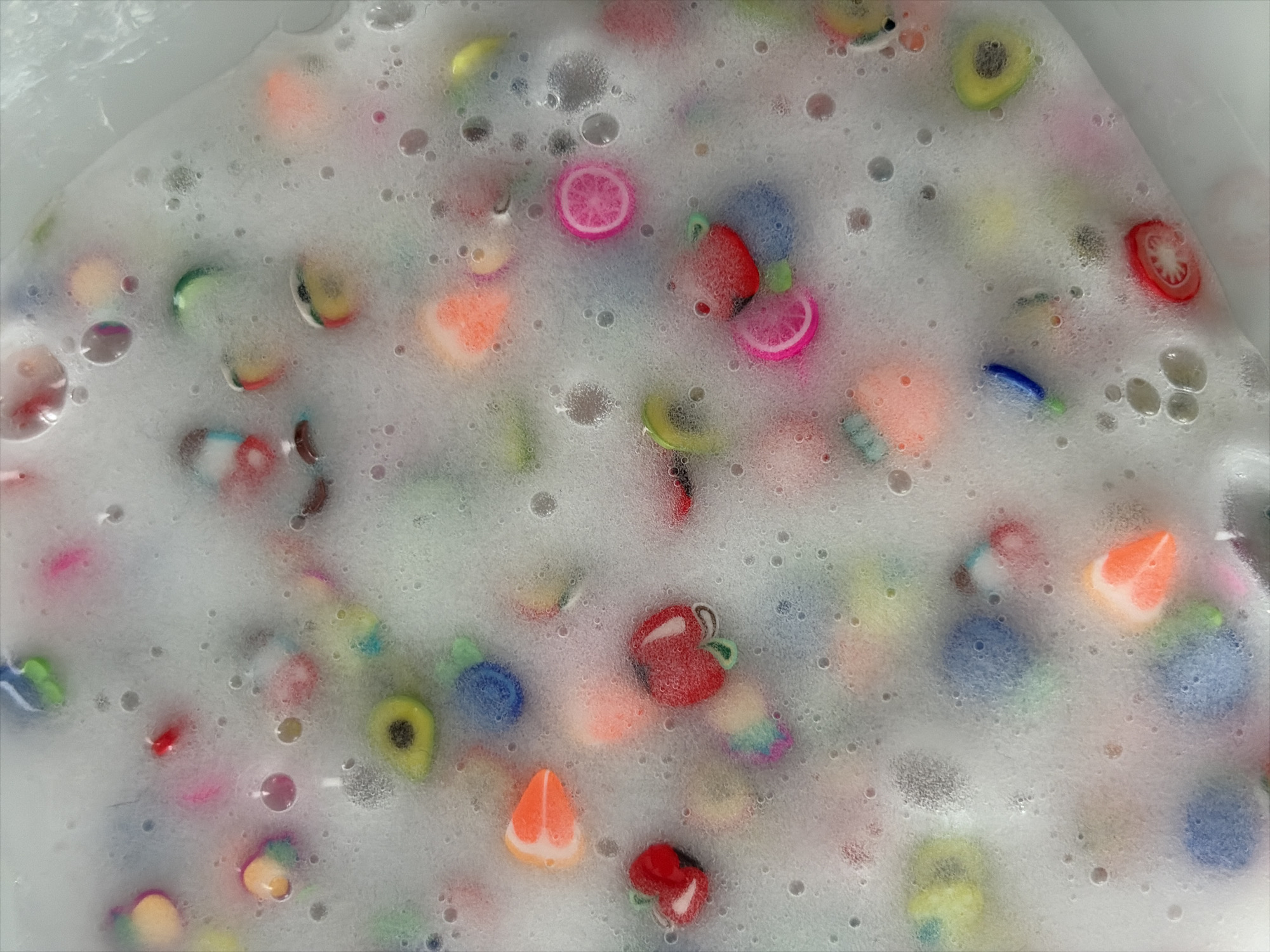 The image is a close-up, overhead shot of a white tub filled with soapy water and numerous colorful, miniature fruit and vegetable charms. The charms are made of polymer clay and include representations of apples, oranges, strawberries, avocados, tomatoes, limes, lemons, and other fruits. They are submerged in the foam, with some floating on the surface and others partially obscured. The water is frothy and bubbly, obscuring the bottom of the tub. The lighting is bright and diffused, creating a soft focus effect. No people or specific Moscow locations are visible.