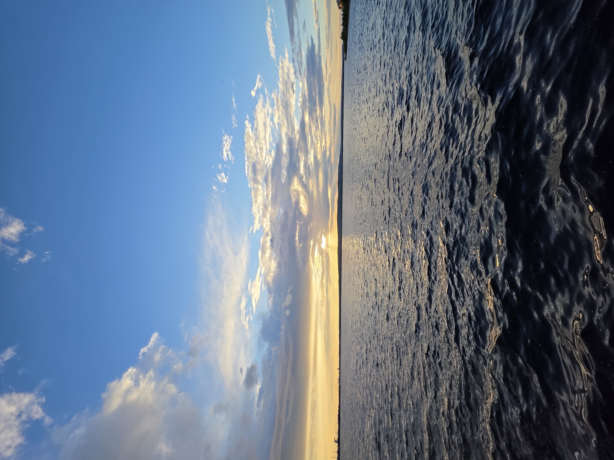 The image is a landscape photograph taken from the water, oriented vertically. The scene depicts a body of water stretching to the horizon, with a sunset sky on the left and rippling water on the right.
The sky is a vibrant blue, dotted with white and gray clouds that are illuminated by the setting sun, creating streaks of yellow and orange light. The sun itself is not directly visible but its glow is apparent along the horizon. The water is dark and reflective, with the sunlight creating shimmering patterns on its surface. The horizon line is sharp, separating the sky from the water and the distant land. Along the distant shoreline, silhouetted trees and structures are visible, suggesting a landmass.
The overall mood is tranquil and serene, characteristic of a sunset over water. The time of day is clearly sunset, indicated by the warm colors in the sky and the sun's low angle. The location is suggested to be Chaikovsky, Russia, implying a natural body of water such as a river or lake in that region. There is no visible text in the image.