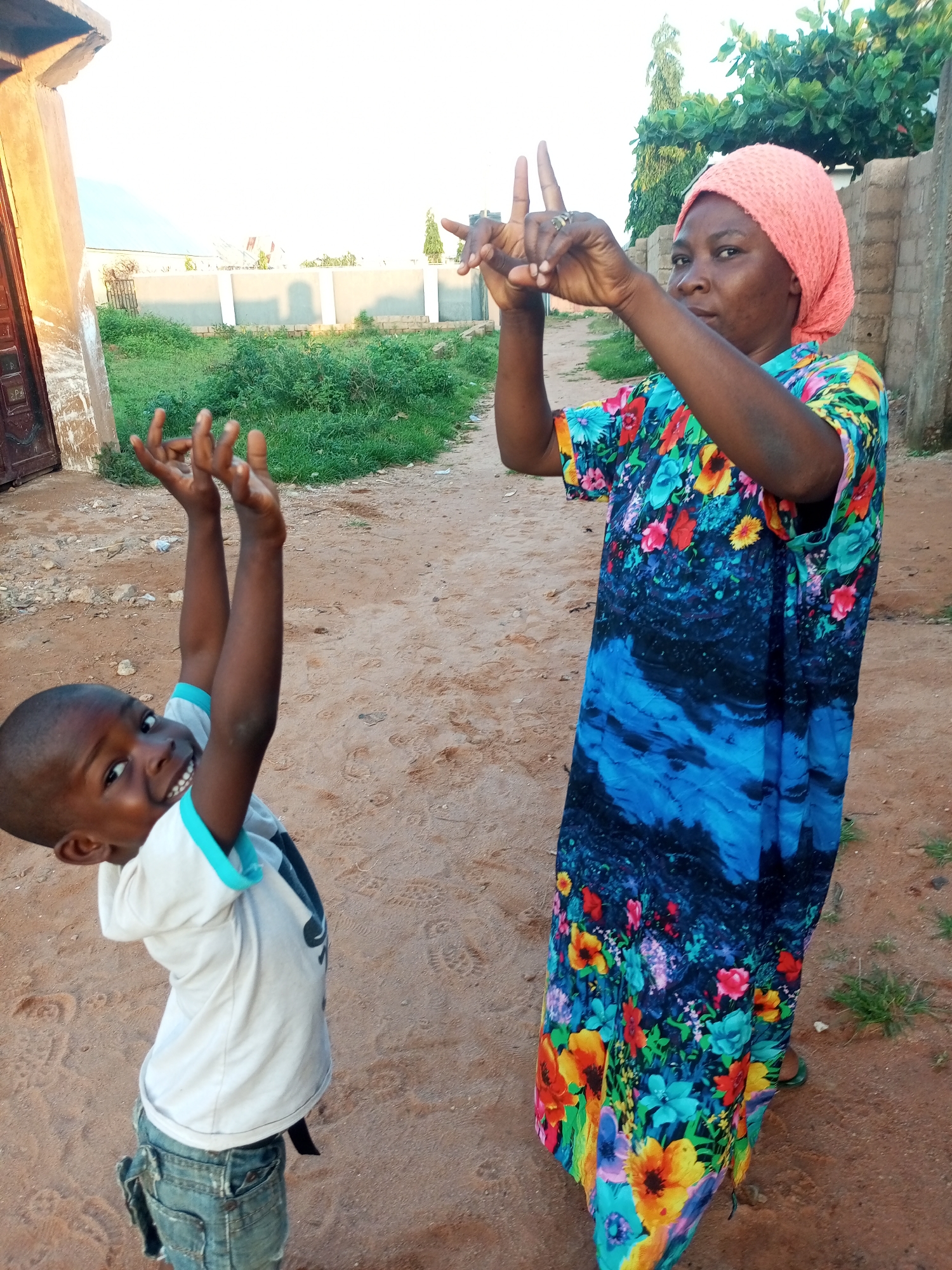 In Tempure, Nigeria, a young boy in a white t-shirt and denim shorts, with his arms raised and hands open, looks up at an adult woman. The woman, wearing a pink headscarf and a vibrantly colored floral dress with a blue abstract pattern, has her hands raised and fingers extended, as if gesturing or signaling. They are standing on a dirt path with sparse vegetation and buildings in the background. The scene appears to be outdoors during daylight.