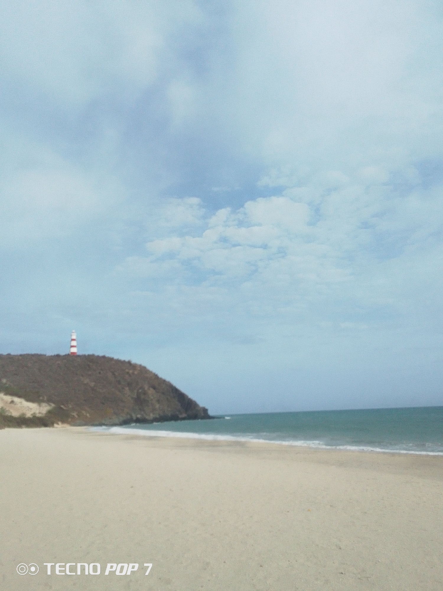 The image depicts a beach in Pedro González, Venezuela. The sky is overcast with white and light blue clouds. The sand is pale and stretches from the bottom of the frame to the water's edge. The sea is a muted teal color with gentle waves lapping the shore. To the left, a landmass rises, topped with a red-and-white striped lighthouse. The vegetation is sparse and brownish. The camera phone brand "TECNO POP 7" is visible at the bottom of the frame. There are no people visible in the image.