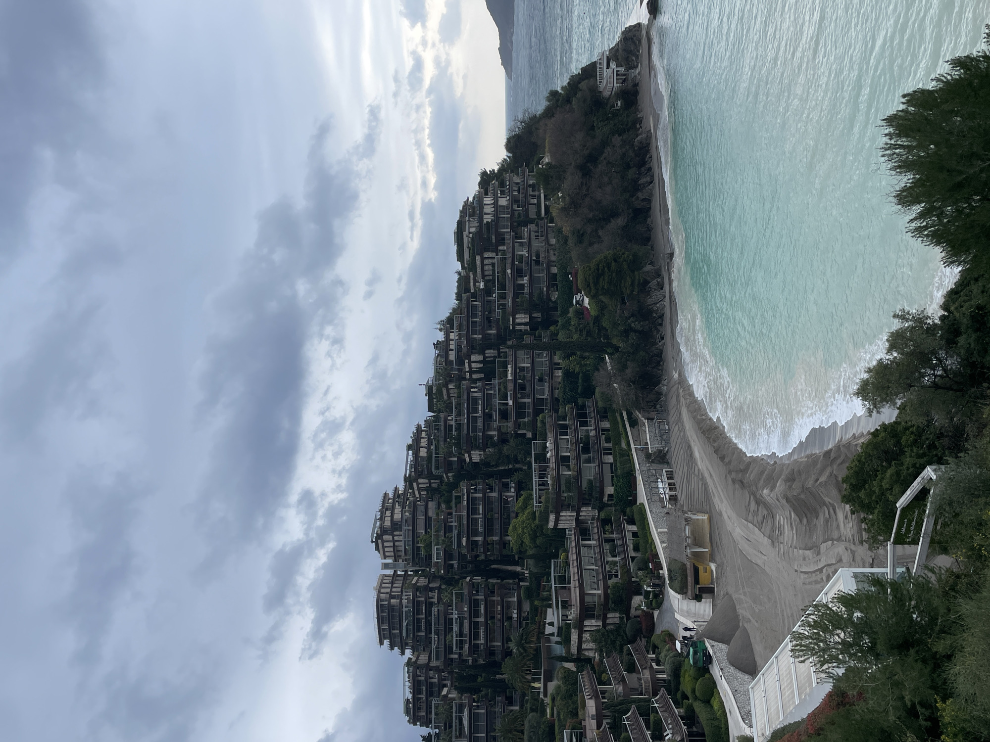 This image captures a coastal landscape on the Zavala Peninsula in Budva, Montenegro, specifically depicting the area of Dukley Gardens and its associated beach. The scene is observed during daylight hours under an overcast sky.
The upper half and central-left of the image are dominated by a complex of multi-story, terraced residential or resort buildings. These structures are integrated into a steep, heavily vegetated hillside, featuring numerous balconies and extensive greenery. Tall, dark green trees and dense shrubs are visible between and around the buildings. A paved road or pathway winds through the lower parts of the complex.
In the midground, a crescent-shaped beach curves from the left towards a rocky promontory on the right. The beach consists of dark grey sand and pebbles, with several piles of sand suggesting recent or ongoing landscaping or re-sanding efforts. Visible tire tracks are present on the flattened beach surface. A small, light-colored utility vehicle, possibly green, is parked on a paved path adjacent to the beach. Two individuals are discernible near this vehicle. Structures consistent with beach facilities, some appearing to be undergoing maintenance or prepared for a season, are scattered along the beachfront. A white sign with dark lettering, reading "Welcome to The Dukley...", is partially visible on a white terraced structure in the lower-left.
The sea, occupying the right and lower portions of the image, displays clear turquoise-blue water near the shore, transitioning to a darker blue further out. Moderate waves with white caps are breaking onto the beach. A rocky, vegetated coastline extends into the sea on the right, culminating in a small, rocky point. In the far distance on the horizon, a darker landmass or island is partially visible.
The sky is entirely overcast with a dense layer of grey clouds, providing diffuse lighting across the scene.