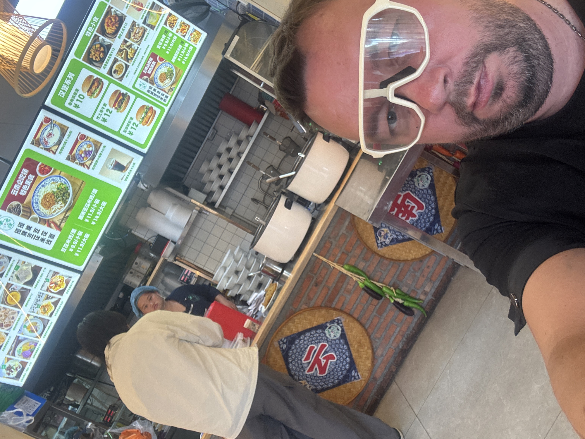 In Suzhou Industrial Park, China, a man wearing white wraparound sunglasses and a black t-shirt is taking a selfie from a seated position. He is positioned in the foreground, with his face and upper chest filling the frame. Behind him, a food stall is visible. Two female employees are working at the counter. One, in the middle distance, wears a blue hairnet and a dark uniform. The other, to her left and further back, wears a white shirt and gray pants, and appears to be serving or interacting with the counter. Above the counter, a digital menu board displays food items and prices in Chinese. The stall has a rustic brick wall, and decorative circular mats with Chinese characters are hung on the wall. Hanging from a string are several green vegetables, possibly eggplants and peppers. Large white pots, likely for cooking, are also visible on the counter. The environment suggests a casual dining establishment.