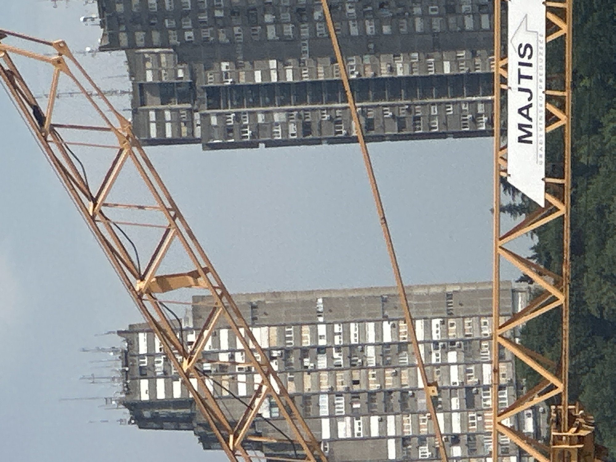 This media file features a construction crane with a yellow lattice boom in the foreground, angled from the bottom left to the top right. Behind the crane, two large, grey apartment buildings are visible. These buildings are characterized by their repeating grid of windows and balconies, suggesting a dense urban environment. The image is a vertical shot, with the buildings appearing somewhat distorted as if viewed from an unusual angle or with a fisheye lens.
On the right side of the crane's boom, a white banner is attached. The banner contains black text in a large, bold font that reads "MAJTIS". Below this, smaller text is present, which appears to be an address or company slogan. An arrow icon is also depicted on the banner, pointing to the right.
The scene appears to be an outdoor setting, likely during daylight hours. The sky behind the buildings is a pale, hazy grey, indicating overcast conditions or a light-filled sky. To the far right, a dense cluster of green trees can be seen, providing a natural contrast to the man-made structures. The overall impression is one of ongoing construction or development in a city. The focus is on the architecture and the construction equipment, with no people visible in the frame. The location is identified as Belgrade, Serbia, which aligns with the style of the apartment buildings and the potential Slavic origin of the text on the banner.