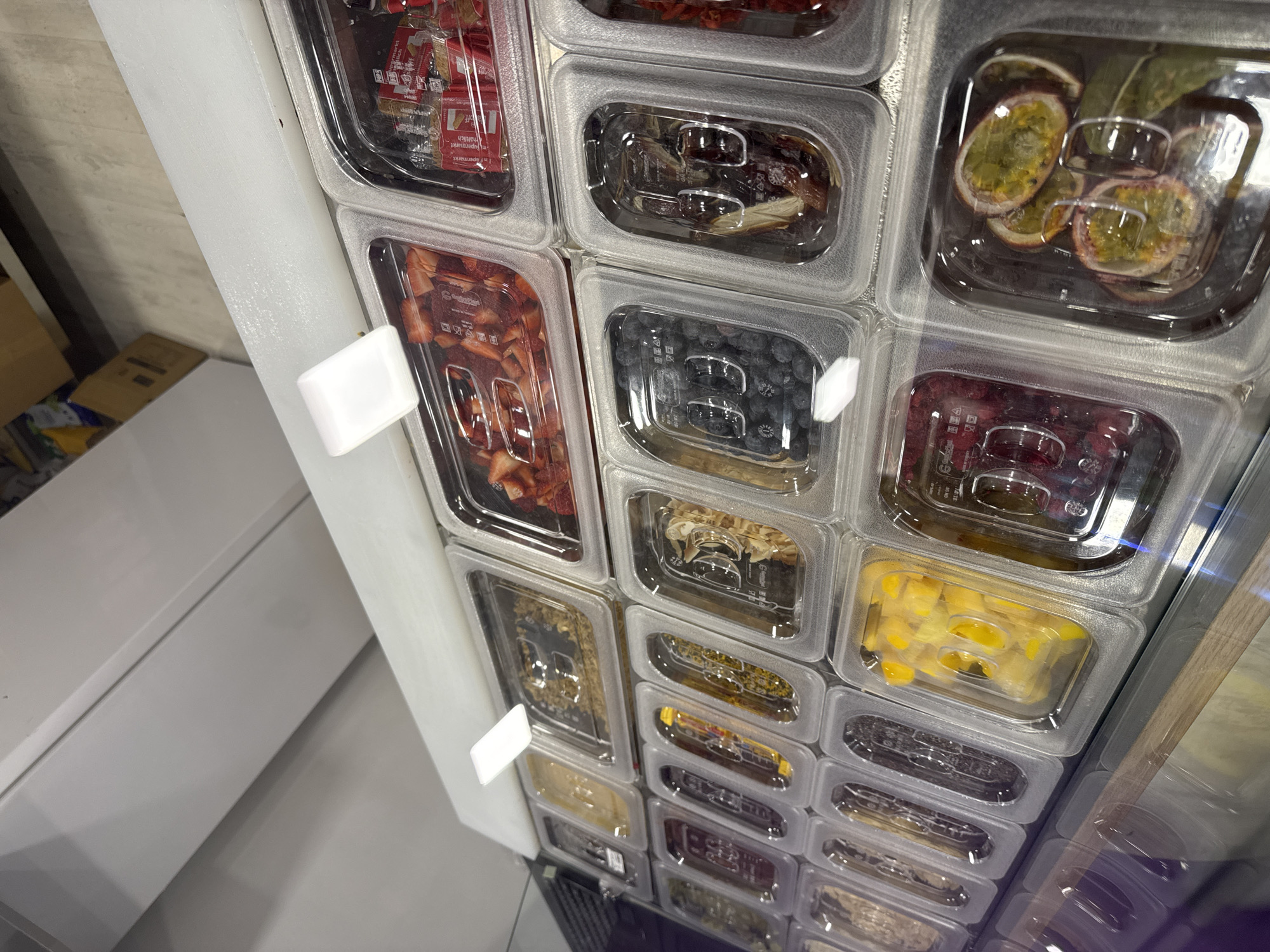 The image shows a vertical display of clear plastic containers filled with various food items. The containers are arranged in a grid pattern, with the contents visible through the transparent lids. Some of the identifiable items include strawberries, blueberries, passion fruit, dried fruits, corn, and a variety of other toppings or ingredients. The display appears to be part of a food preparation area, possibly a salad bar or yogurt bar, where customers can select toppings. White plastic tags are attached to the side of the display. The environment is clean and well-lit. The specific location within Sofia, Bulgaria, cannot be determined from the image alone. There are no people visible in the scene.
