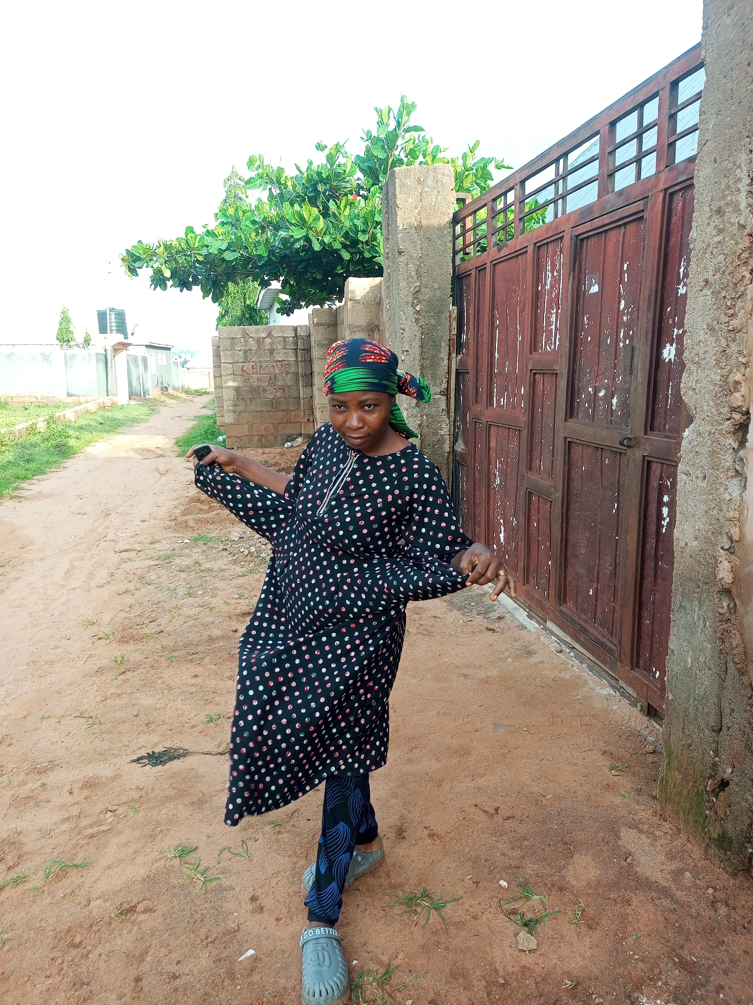 In Tempure, Nigeria, a woman stands on a dirt path in front of a rusty, weathered metal gate. She is wearing a black dress with a white and pink polka dot pattern and a green, black, and red headscarf. Her left hand is extended, fingers splayed, and her right hand is partially visible. She wears blue and black patterned leggings and grey slip-on shoes. The background features a stone wall, some vegetation including a tree with broad leaves, and a distant view of buildings and a water tank. The sky is bright and overcast.