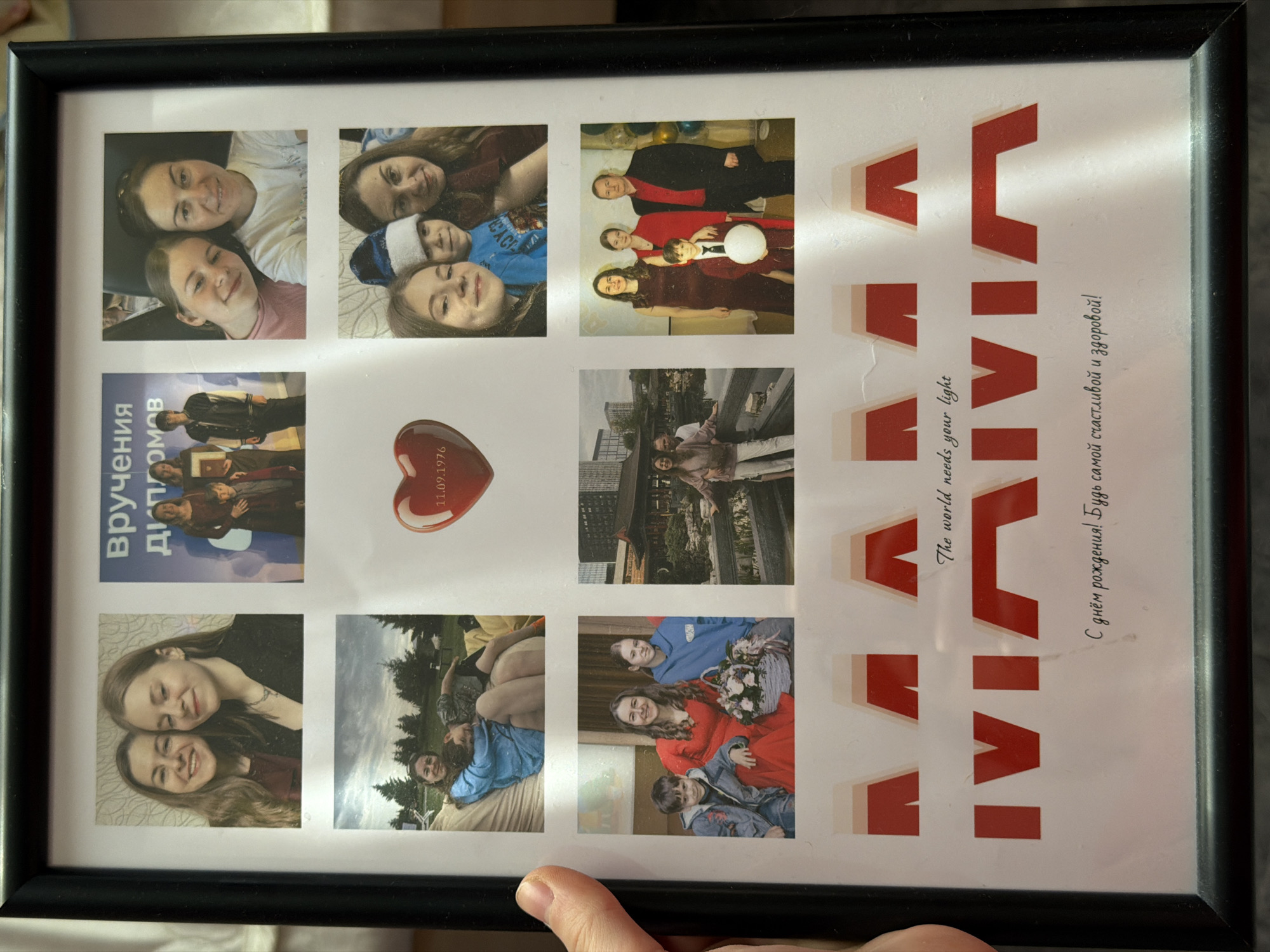 A framed photo collage, oriented vertically, displays a collection of personal photographs, seemingly dedicated to an individual named Vivian. The black frame encloses a grid of eleven smaller images, a red heart, and several lines of text in both Russian and English.
The photographs feature various individuals, primarily women and children, often smiling and posing together. They capture different moments and settings: some are indoors, possibly at social gatherings or formal events, while others are outdoors amidst natural scenery or urban backdrops. One prominent photo shows a group, including women in academic gowns, with vertical text alongside it reading "Вручения Дипломов" (Diploma Ceremony), indicating a graduation event. Other photos depict family or friends celebrating, with elements like balloons and a basket of flowers visible in different images.
A red heart-shaped object in the center column bears the date "11.09.1976," likely a significant birth date or anniversary. The right side of the collage features large, red, stylized letters spelling out the name "VIVIAN." Below this name, in English, is the phrase "The world needs your light." Further down, in Russian, it reads "С днём рождения! Будь самой счастливой и здоровой!" (Happy Birthday! Be the happiest and healthiest!). This text strongly suggests the collage is a birthday tribute to Vivian. The overall mood conveyed by the images is joyful and celebratory, showcasing cherished memories in Perm, Russia.