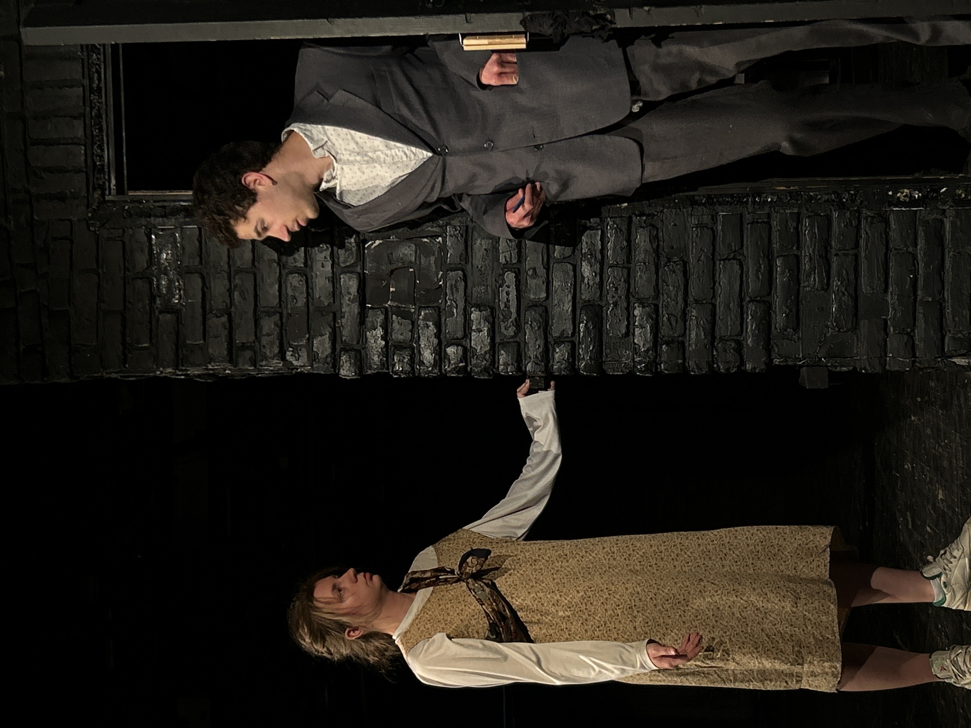 A medium shot depicts two individuals. The top individual, a male, is dressed in a grey suit and a white patterned shirt, leaning out of a black brick window, looking down. He holds a pen in his left hand and a notepad in his right. Below, a female in a beige dress over a white long-sleeved shirt reaches upwards with her right hand towards the man. The setting appears to be a theatrical stage. The city and specific location are not discernible.