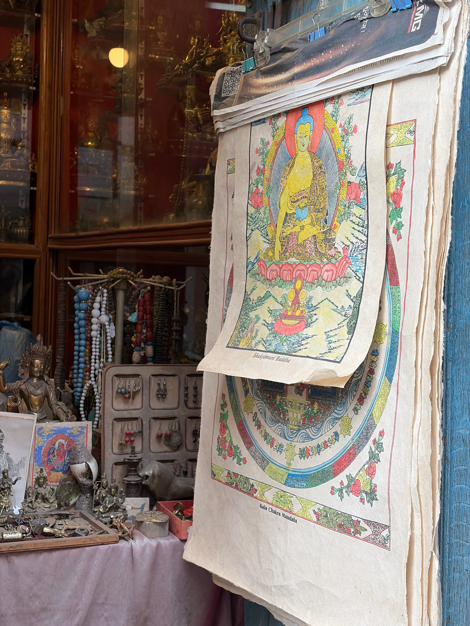 In Lalitpur, Nepal, an outdoor market stall displays a collection of Tibetan Buddhist art and religious items. In the foreground, a stack of unframed paintings on textured paper leans against a blue wooden surface. The topmost painting features a depiction of Shakyamuni Buddha, seated in meditation with a yellow halo and robes, surrounded by a vibrant floral border. Below it, another painting displays a Kala Chakra Mandala, a complex circular diagram with intricate patterns and Buddhist symbols.
To the left of the paintings, a variety of religious paraphernalia is visible. A bronze statue of a multi-armed deity stands prominently. Arranged on a rack are numerous strands of prayer beads in various colors, including blue, white, and red. Below the beads, a display case holds an assortment of small earrings and other trinkets. Further to the left, shelves within a glass-fronted cabinet contain numerous smaller golden statues and religious artifacts, illuminated by a light source within the cabinet. A pink tablecloth covers the surface of the stall. The overall scene suggests a vibrant and spiritual marketplace setting.