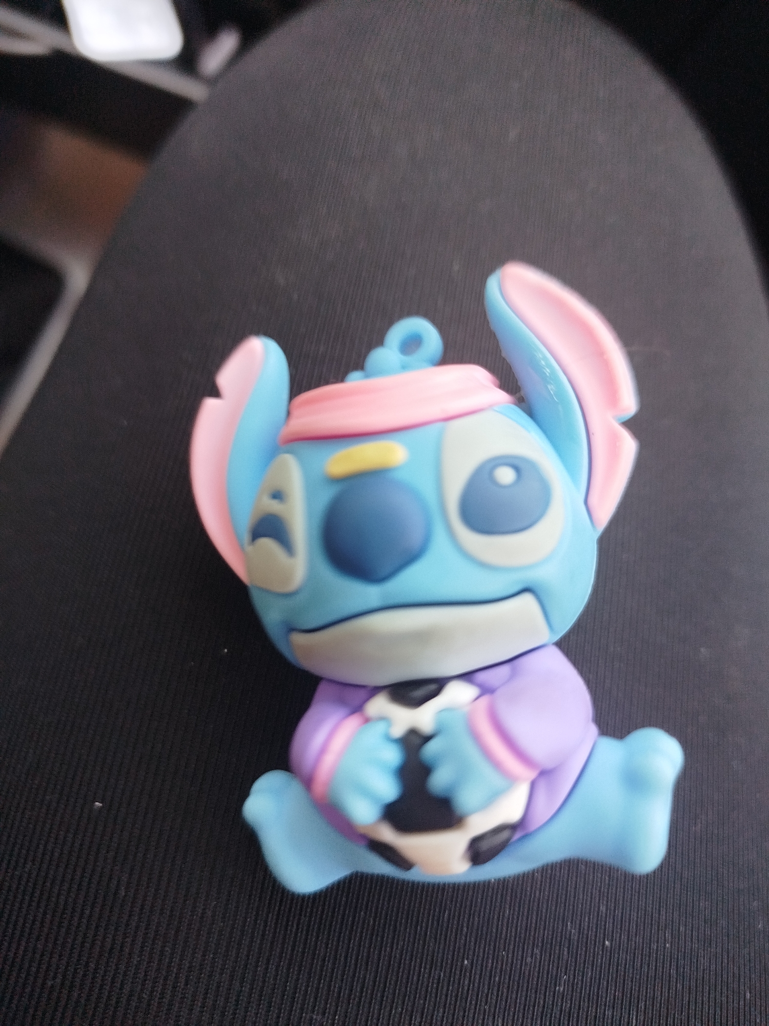 This media file contains a close-up, overhead shot of a small, blue, Stitch-like toy figure sitting down and holding a black and white soccer ball. The figure has large, pointed ears with pink inner linings, a pink band around its head, and a blue face with yellow markings over its eyes. It is wearing a purple outfit with pink trim. The figure's hands are wrapped around the soccer ball. The toy is placed on a dark, textured surface, possibly fabric. The lighting suggests it is indoors, with a soft light source coming from above and to the left. There is no visible text in the image, and the overall mood is playful and cute.