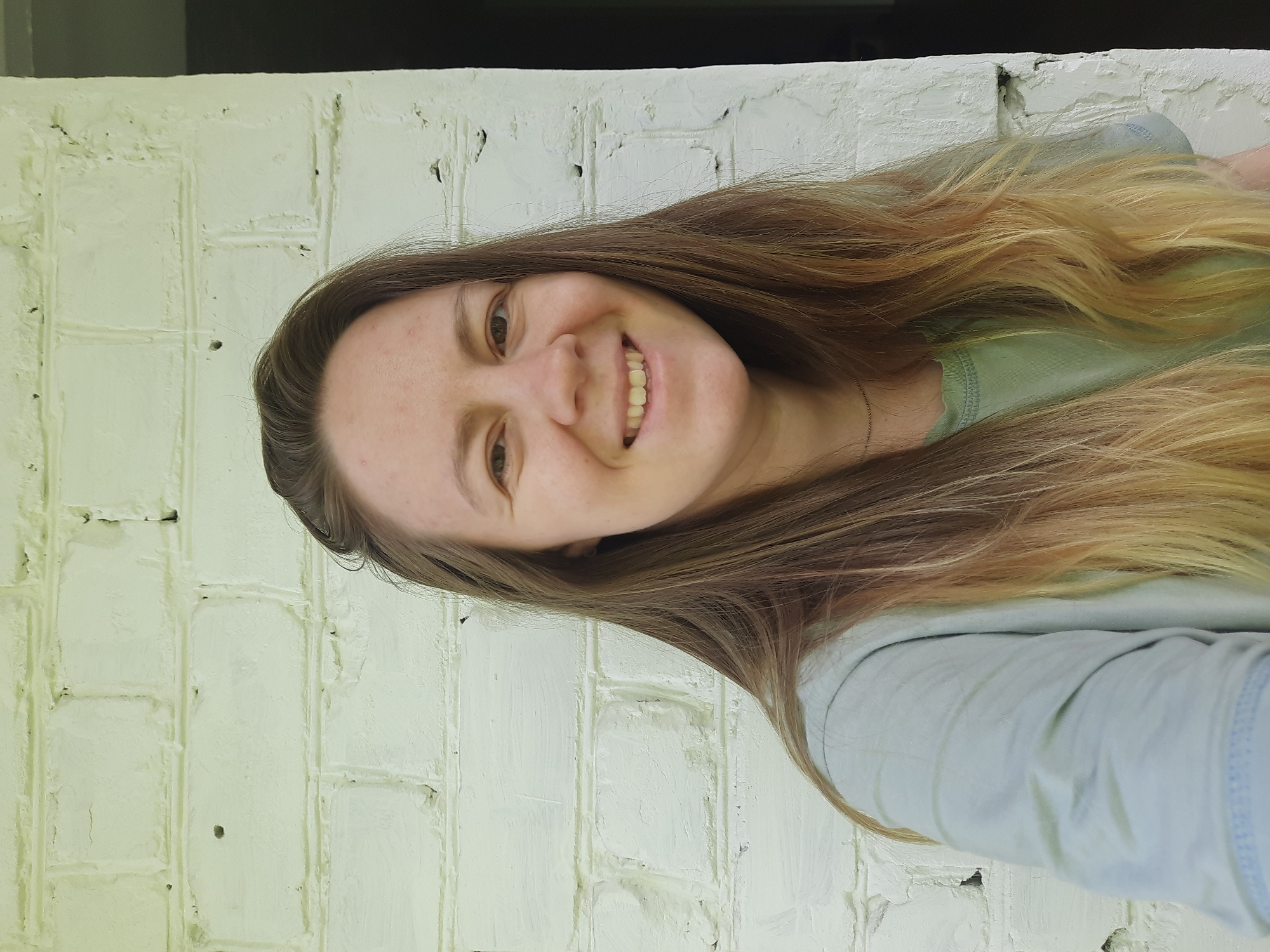 A young woman with long, light brown hair is smiling in front of a white-painted brick wall. Her hair has blonde highlights toward the ends. She is wearing a light green t-shirt. The background is the white brick wall. The location cannot be determined based solely on this image, but based on the user's prompt, it is located in the city of Izhevsk, Russia.