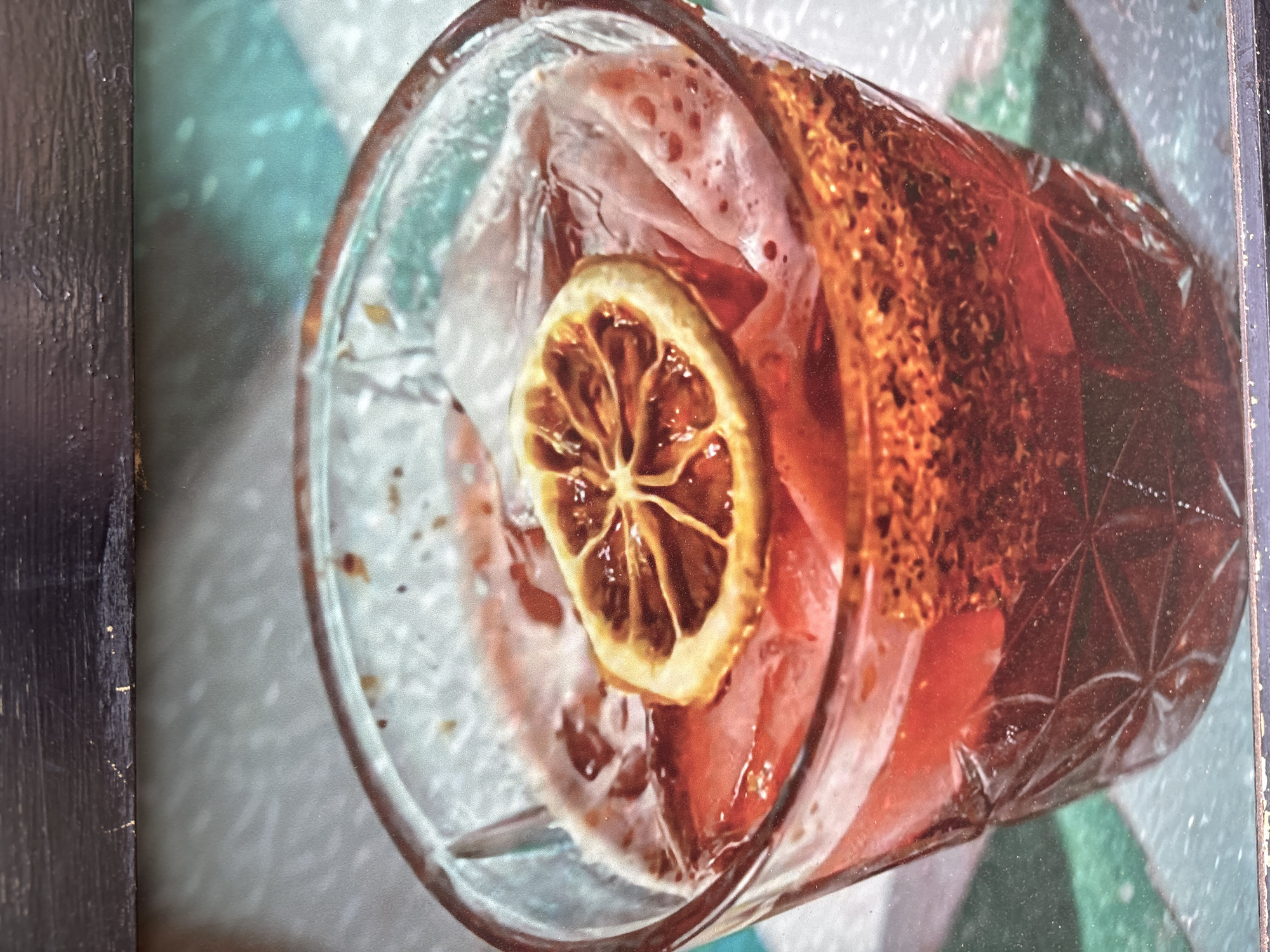 This is a close-up, top-down shot of a cocktail in a clear, faceted glass. The drink is a deep reddish-brown color and appears to be chilled, as evidenced by the crushed ice and condensation on the glass. A single, dried lemon slice rests on top of the ice, its segments clearly defined. The drink is garnished with what appears to be a thick, textured rim of spices or sugar, which is a golden-brown color. The background is out of focus but suggests a patterned surface, possibly a tablecloth or countertop, with teal and white elements. The lighting is soft, and the overall impression is one of a refreshing and perhaps slightly exotic beverage, likely served in a bar or at a casual gathering. There is no text visible in the image.