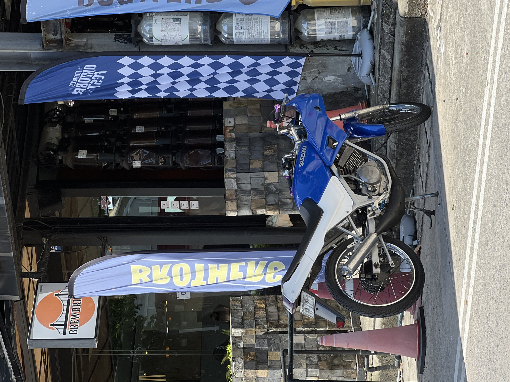 A blue and white Suzuki motorcycle is parked on a sidewalk in front of a building with large glass windows. The motorcycle is leaning on its kickstand. A tall orange traffic cone is to the left of the motorcycle. Two tall, blue, feathered flags are planted on the sidewalk, one to the left and one to the right of the motorcycle. The flag on the left has yellow text that reads "BROTHERHOOD" in large letters. The flag on the right has a checkered pattern and text that is difficult to read. Behind the motorcycle, through the glass windows of the building, a stack of large, dark glass containers can be seen. A white sign with an orange circle containing a bridge and the text "BREWBRIDGE" is mounted above the entrance to the building. The ground is paved with concrete and asphalt, with white painted lines indicating a road.