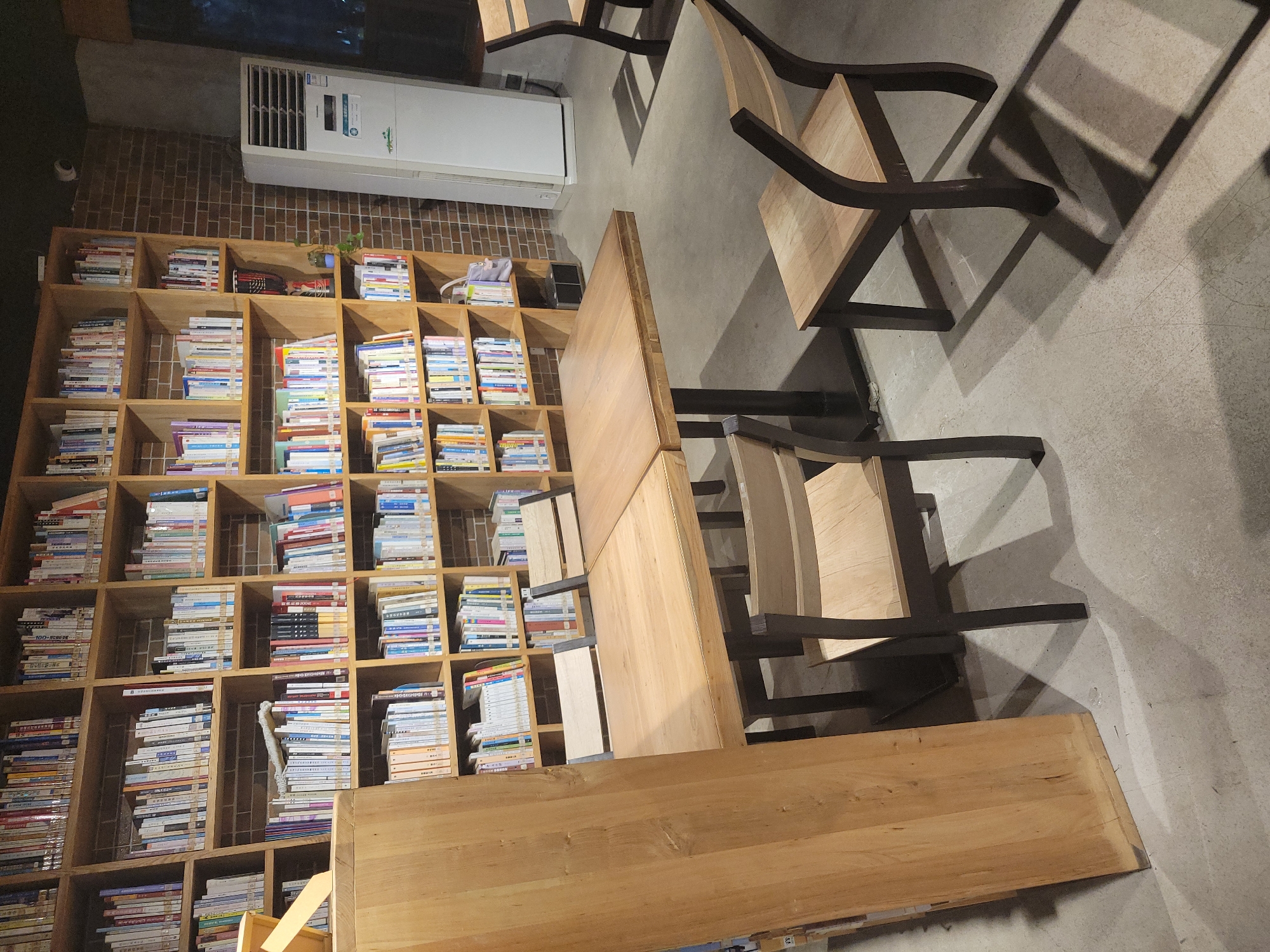 The image shows an interior scene in what appears to be a library or study area. In the background, there is a large wooden bookshelf filled with books. To the right of the bookshelf, there is a white air conditioning unit. In the foreground, there are wooden tables and chairs with dark-colored frames. The floor is a light gray color. The location is Shapingba District, China.