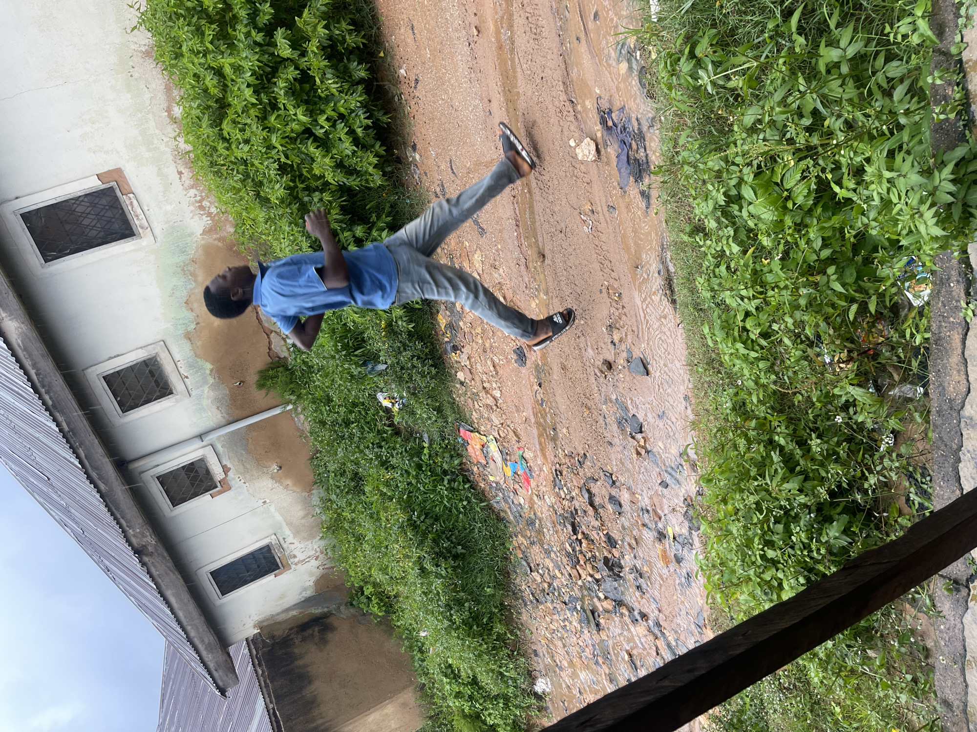 A young Black male, wearing a blue short-sleeved shirt and grey pants, is captured mid-stride, running on a muddy, debris-strewn path. The path appears to be a road or an open area with standing water in places. To the left of the path is a weathered, light-colored building with a corrugated metal roof and several windows. The building's facade shows signs of age and damage, with exposed brickwork and peeling paint. Dense green foliage lines the path and surrounds the building, with some scattered litter visible amongst the vegetation and on the path itself. The environment suggests an outdoor, possibly rural or semi-urban setting. The overall impression is one of overcast weather, as evidenced by the diffuse lighting. The location is the city of Rafin Par, Nigeria.