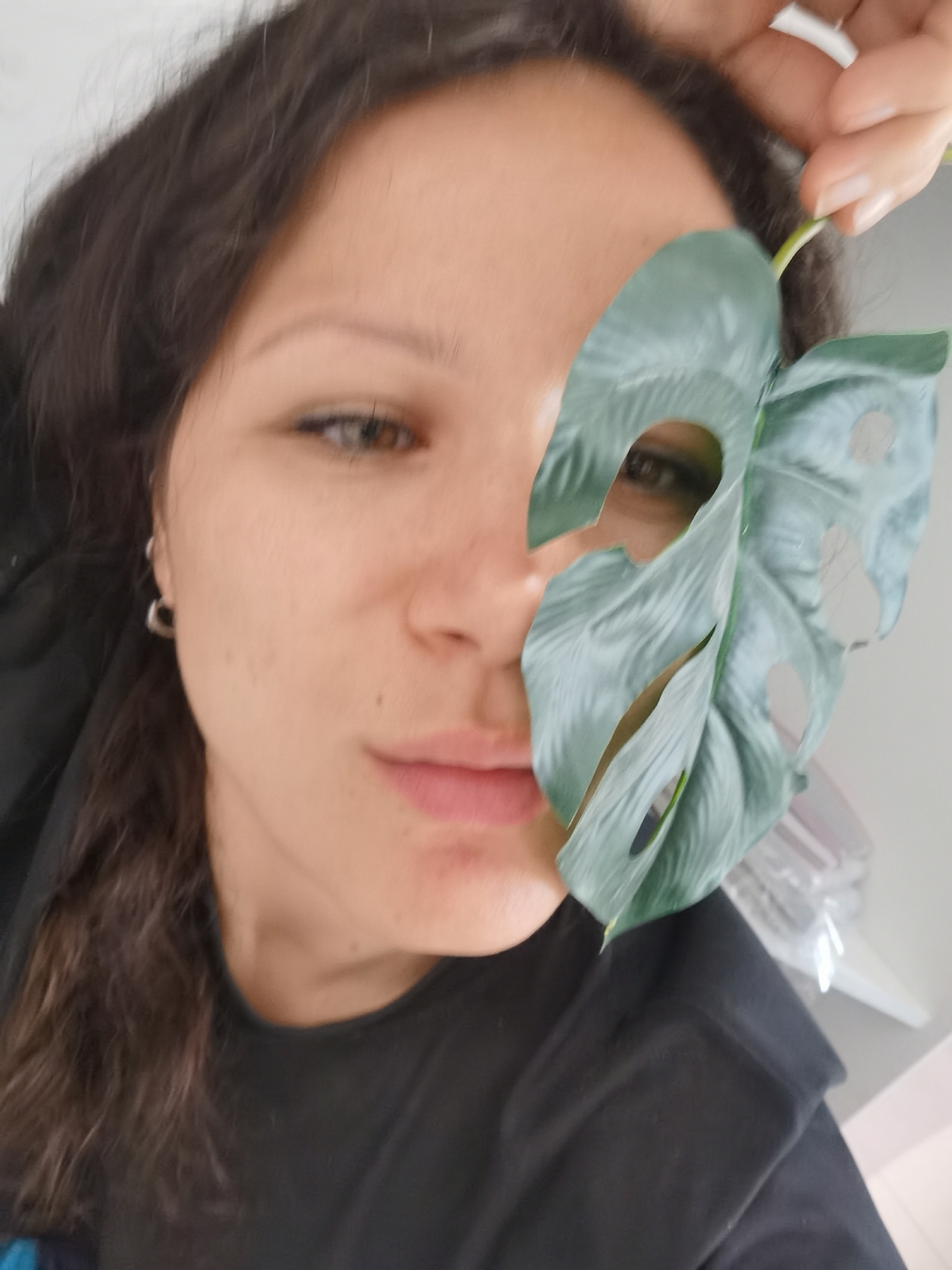 A woman poses for a selfie, partially obscured by a split-leaf philodendron leaf held in front of her face. She has dark, curly hair and is wearing a black shirt. Her skin tone is light. A small hoop earring is visible in her left ear. The environment is blurred and appears to be indoors. The image location is not verifiable as Chaikovsky, Russia based on the content.