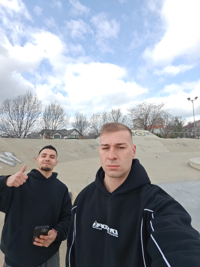 Two adult males are photographed in a selfie-style shot within Skate Park Shumen in Shumen, Bulgaria. The male positioned closer to the camera, on the right, has short light hair and a serious expression, wearing a black hooded sweatshirt with white piping along the arms and sides, and a white graphic logo (stylized 'DROP') on the chest. A silver chain is visible at his neck. He is looking directly forward, presumably holding the camera. The second male, situated behind and to the left, has dark hair, a dark goatee, and a smiling expression. He wears a plain black hooded sweatshirt, holds a black smartphone in his left hand, and displays a thumbs-up gesture with his right hand. The background consists of various concrete skate park obstacles, including ramps and a central pyramid-like structure with visible graffiti. Beyond the park structures, leafless deciduous trees line the scene, with residential houses and a taller apartment building visible behind them. A tall metal street light with multiple lamps is present on the far right. The sky overhead is partly cloudy, displaying patches of blue amidst white and grey clouds.