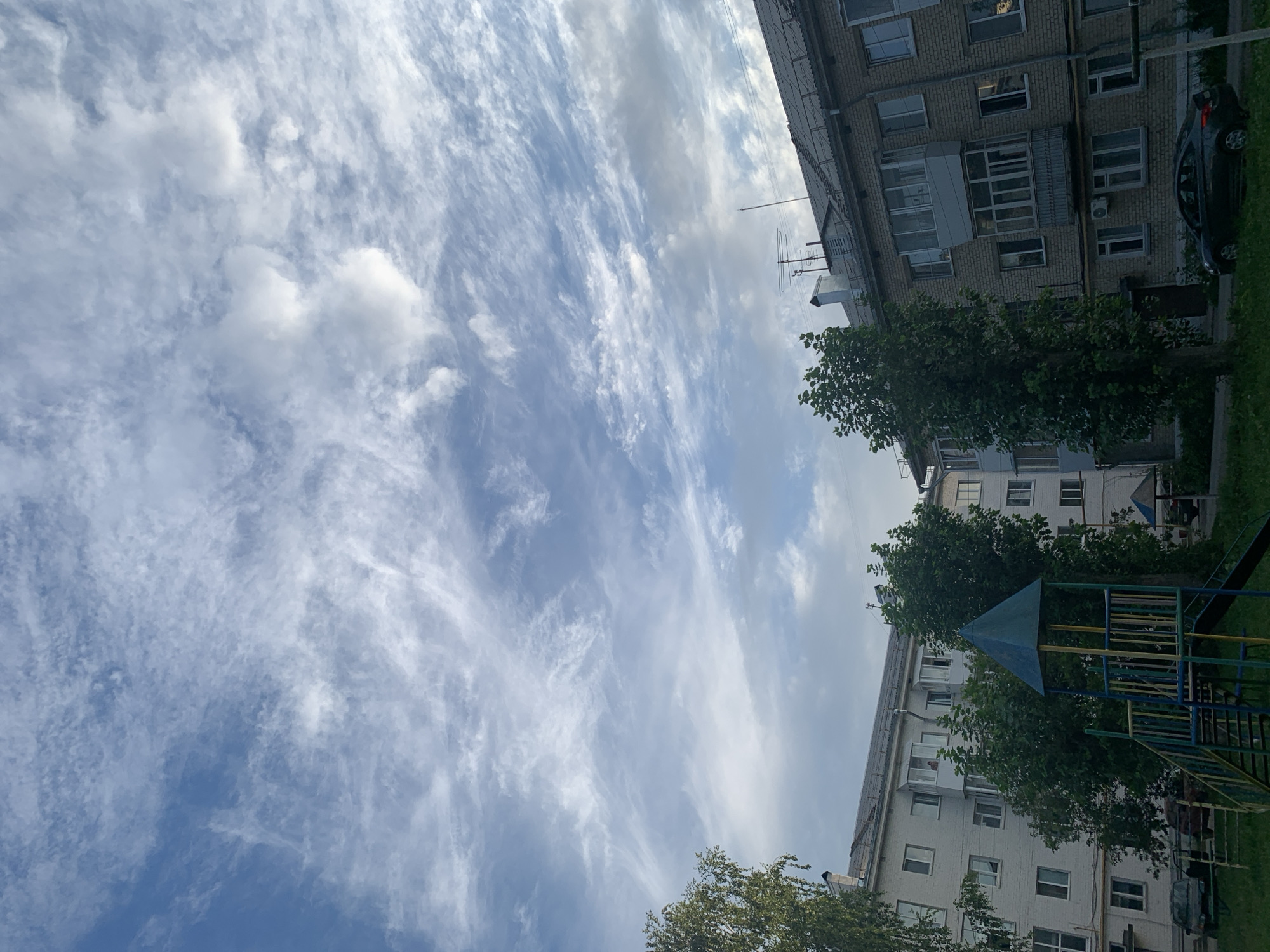 This image depicts a residential area in Asha, Russia, under a sky filled with wispy cirrus clouds. In the foreground, a children's playground structure with a blue conical roof and yellow and blue railings is visible on a grassy area. Behind the playground are two apartment buildings. The building on the left is lighter in color and appears to have a simpler facade, while the building on the right is constructed of brick and features balconies and satellite dishes on the roof. Several cars are parked along the street in front of the buildings. The overall impression is a typical urban scene on a cloudy day.