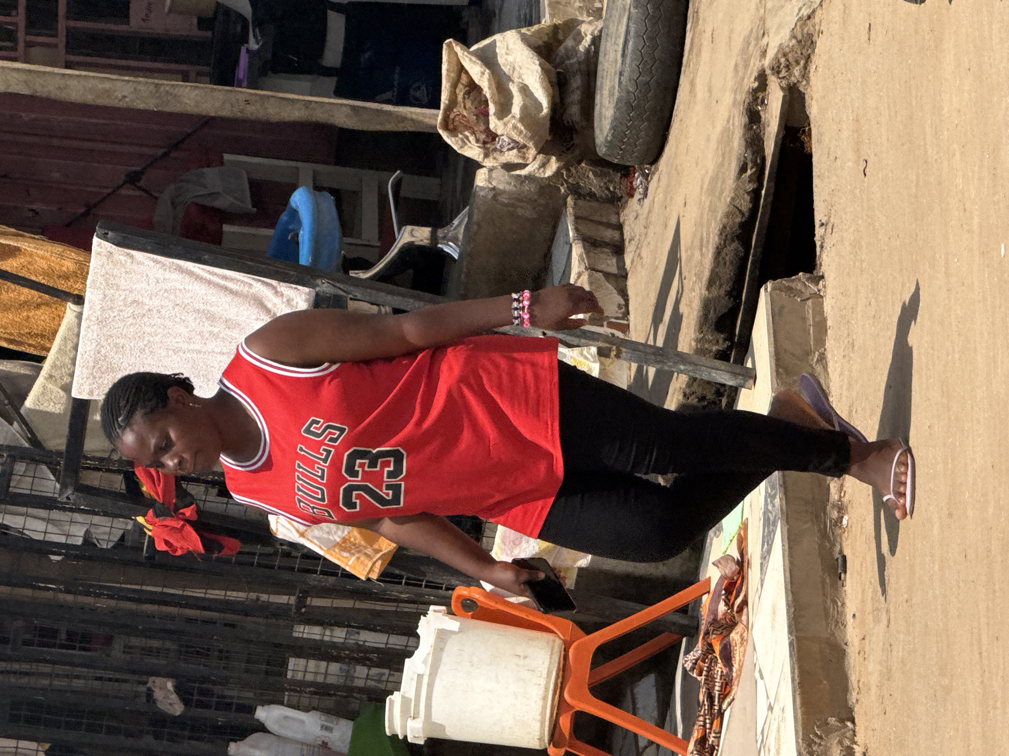A woman wearing a red basketball jersey with "Bulls 23" on it, black leggings, and flip-flops walks down a dirt path in Abuja, Nigeria. She is holding a dark-colored mobile phone in her left hand. To her left, a large white plastic container sits on an orange chair. In the background, there are various items, including fabrics, a blue plastic basin, a sack of contents, and a tire. The environment appears to be an outdoor marketplace or a residential area with a mix of structures and materials.