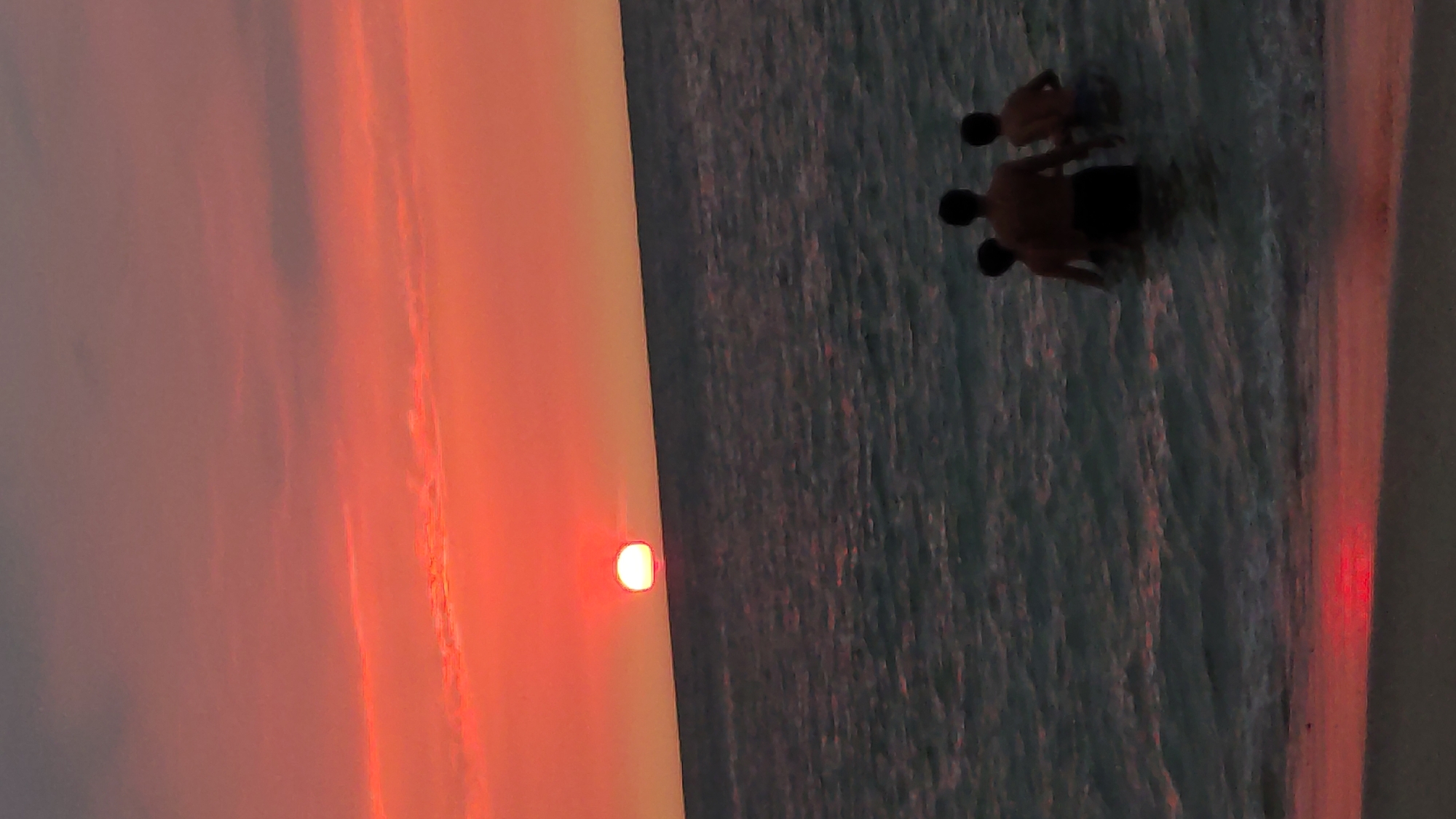 The image shows a sunset over the ocean in Bocapán, Peru. Three people are in the water near the shore. The sky is a gradient of orange and pink, with the setting sun visible on the horizon.