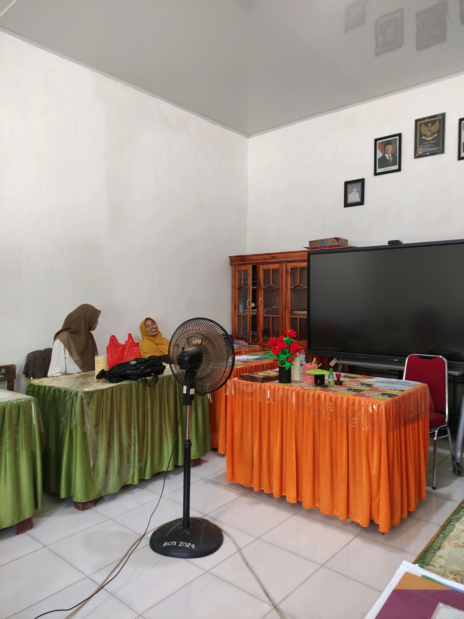 The image depicts an indoor scene, likely a classroom or meeting room in Bulukumba, Indonesia. The room is furnished with several tables draped in green and orange cloths. A black oscillating fan stands in the foreground, its base marked with "BOS 2024." Two women are seated at a table covered with a green cloth. One woman, wearing a brown hijab, faces away from the camera, while the other, in a yellow hijab, is laughing and looking towards the right. Red plastic bags and black bags are on the table with them.
Behind them, a wooden cabinet with glass doors is visible, filled with various items. To the right of the cabinet, a large black screen or television is mounted on a stand. A red chair is positioned in front of this screen. The walls are painted white, and framed pictures of what appear to be official portraits are hung on the wall. The floor is tiled with light-colored squares. The overall impression is of a functional space, possibly used for educational or administrative purposes.