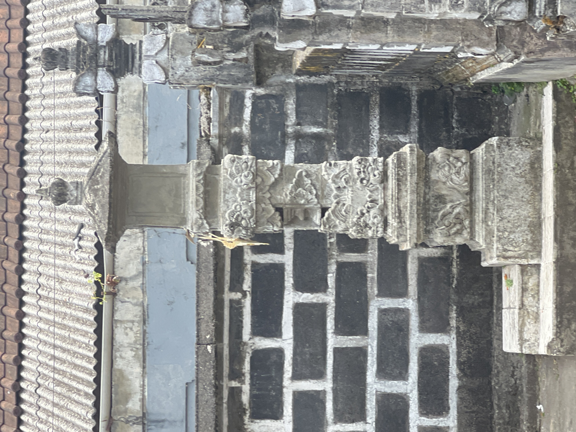 The image shows a stone structure, likely a shrine or monument, located in the city of Denpasar, Indonesia. The structure is intricately carved with decorative motifs, suggesting a cultural or religious significance. It is made of grey stone and appears weathered, indicating its age and exposure to the elements. The background consists of a brick wall and a tiled roof, characteristic of Indonesian architecture. The lighting suggests it is daytime, possibly overcast, as there are no strong shadows. There are no people or animals visible in the frame. The scene appears calm and serene, with a focus on the architectural details of the stone structure.