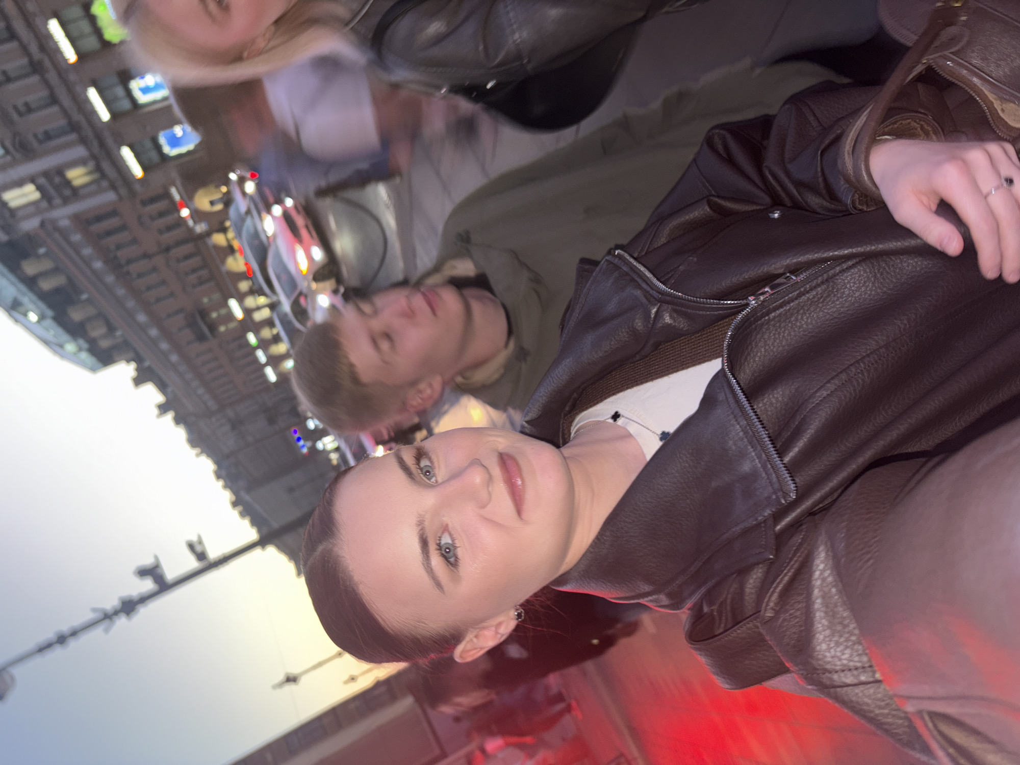 A group of young adults are seen on a street at dusk in Saint Petersburg, Russia. The lighting suggests it is either sunrise or sunset, casting a warm glow on the scene. The main subject, a young woman in the foreground, is looking at the camera with a slight smile. She is wearing a dark brown leather jacket over a white shirt, and a necklace with a dark pendant. Her hair is pulled back neatly. Beside her and slightly behind is a young man in a green hooded jacket. Further in the background, other individuals and vehicles are visible. The street is lined with buildings with ornate architecture, typical of Saint Petersburg. Cars with illuminated headlights are on the road, indicating active traffic. Red lighting is present on the left side of the image, possibly from a sign or decorative lighting. The overall atmosphere appears to be casual and social.