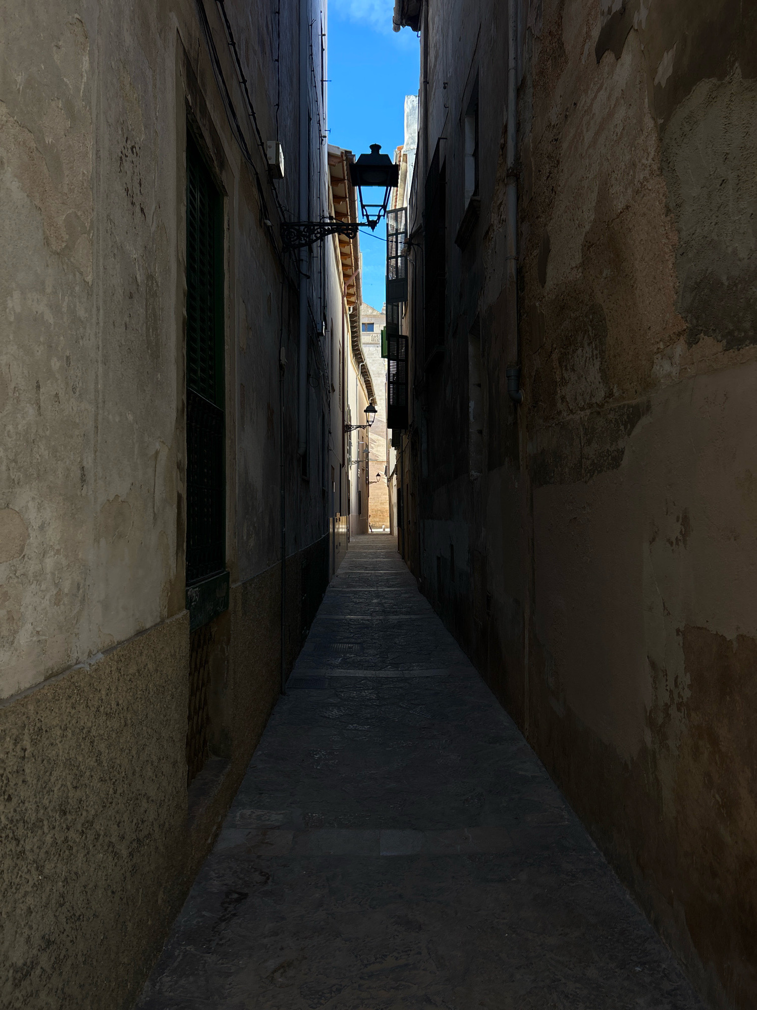 The image depicts a narrow, ancient alleyway in the city of Palma de Mallorca, Spain. The alley is characterized by tall, weathered stone buildings that rise on either side, creating a sense of enclosure. The walls exhibit signs of age, with textured surfaces and peeling paint in shades of beige and brown.
The alley floor is paved with cobblestones, appearing dark and somewhat wet, suggesting recent rain or the natural patina of aged stone. Sunlight casts dramatic shadows, illuminating parts of the path while leaving others in deep shade. Several wrought-iron lampposts are visible, one larger one with a decorative bracket attached to the left building, and a smaller one further down the alley on the right. Some windows with dark green shutters are present on the left side of the alley.
In the distance, the alley opens up slightly, revealing a glimpse of more buildings and a bright blue sky. The architecture suggests a historic Mediterranean setting. No people are clearly visible in the frame, adding to the quiet, timeless atmosphere of the scene. The overall impression is one of a preserved, charming, and historic urban passage.