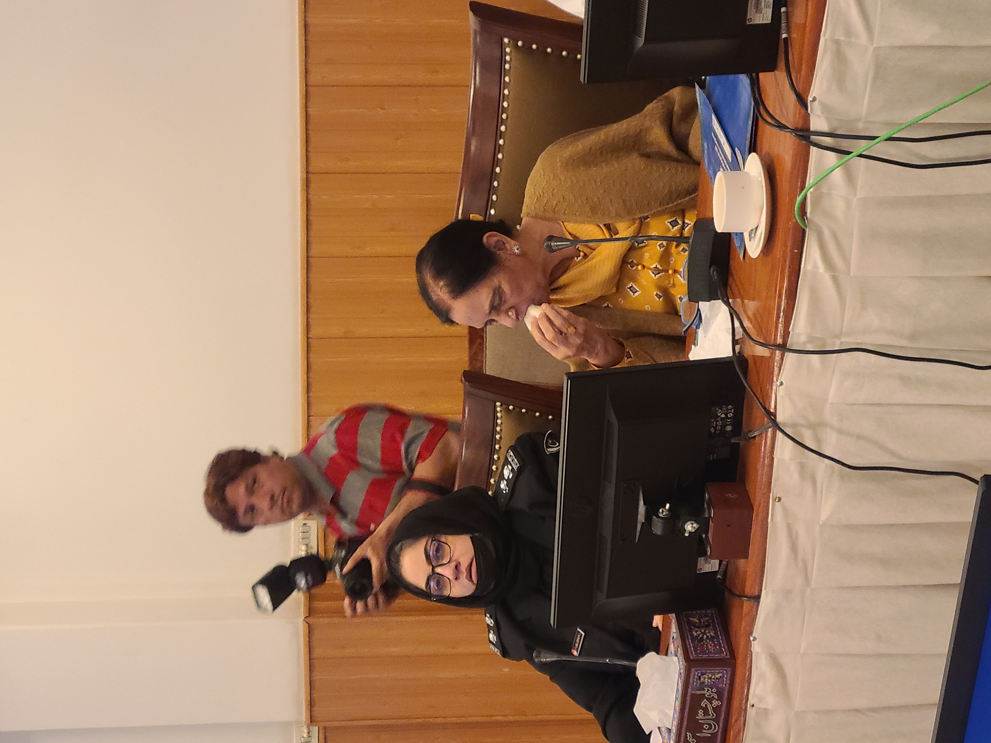 The image shows three people in what appears to be an official setting. A woman in a yellow patterned top and brown shawl is seated and eating something. Behind her, a man with a camera is taking a photograph. In the foreground, a woman wearing a black uniform and a headscarf is seated in front of a computer monitor. The setting includes wooden paneling, furniture, and electronic equipment. The image does not provide enough information to confirm its location in Quetta City Tehsil, Pakistan.