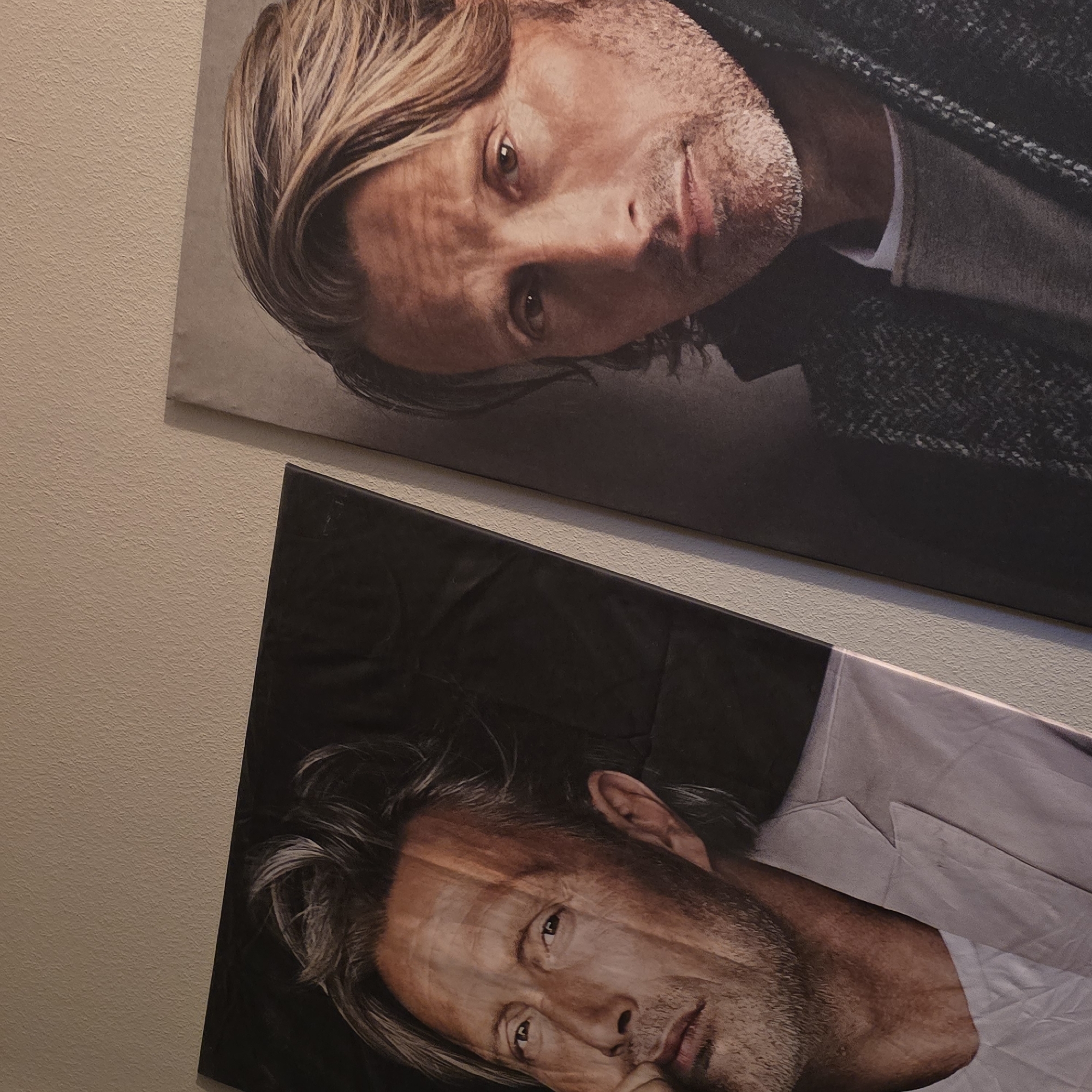 Two large portraits of the actor Mads Mikkelsen are displayed on a textured, light-colored wall. The portrait on the left shows Mikkelsen from the chest up, with his head turned and his hand resting near his mouth. He is wearing a light-colored suit jacket over a white t-shirt. The background of this portrait is dark and appears to be fabric. The portrait on the right is a closer shot of Mikkelsen's face and upper torso. He is looking directly at the camera and is wearing a dark, textured jacket over a grey sweater. The lighting suggests an indoor setting. No specific location in Kettig, Germany, is discernible from the image.