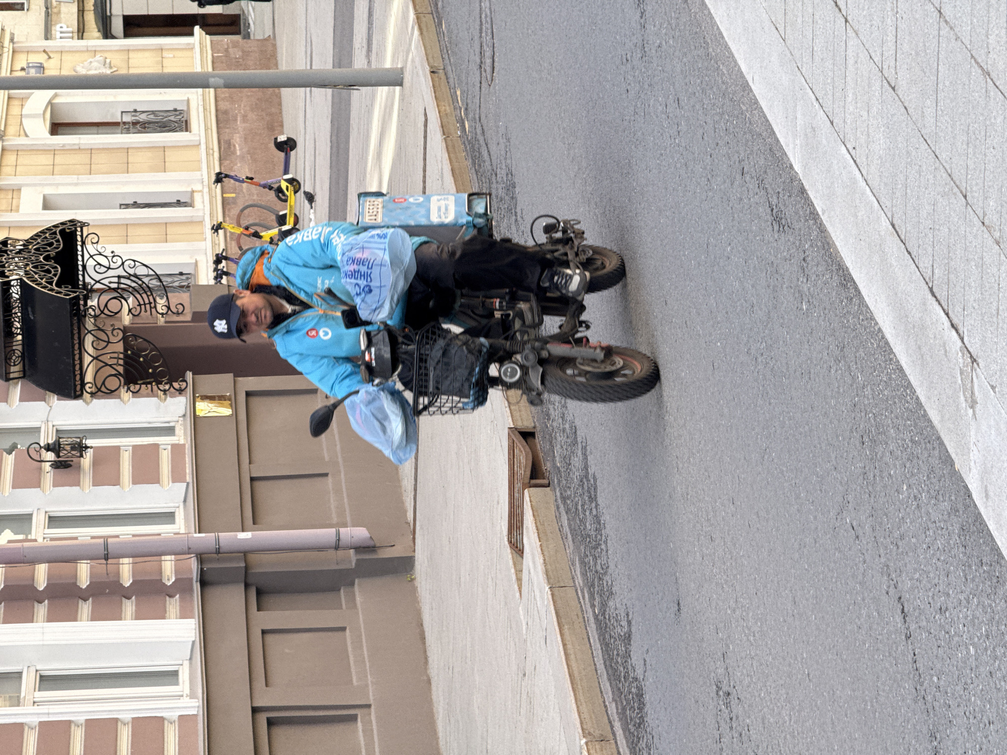 A delivery worker is on a small motorbike on a wet street in Moscow, Russia. He is wearing a light blue jacket with the Yandex logo, a black baseball cap, and dark pants. He is facing left and looking slightly toward the camera. There are blue plastic bags attached to the front of the bike. A delivery box is attached to the back of the bike. The bike is positioned on a wet asphalt street next to a concrete sidewalk. In the background, there is a beige building with wrought iron details and various bicycles. There is a metal pole in the background.