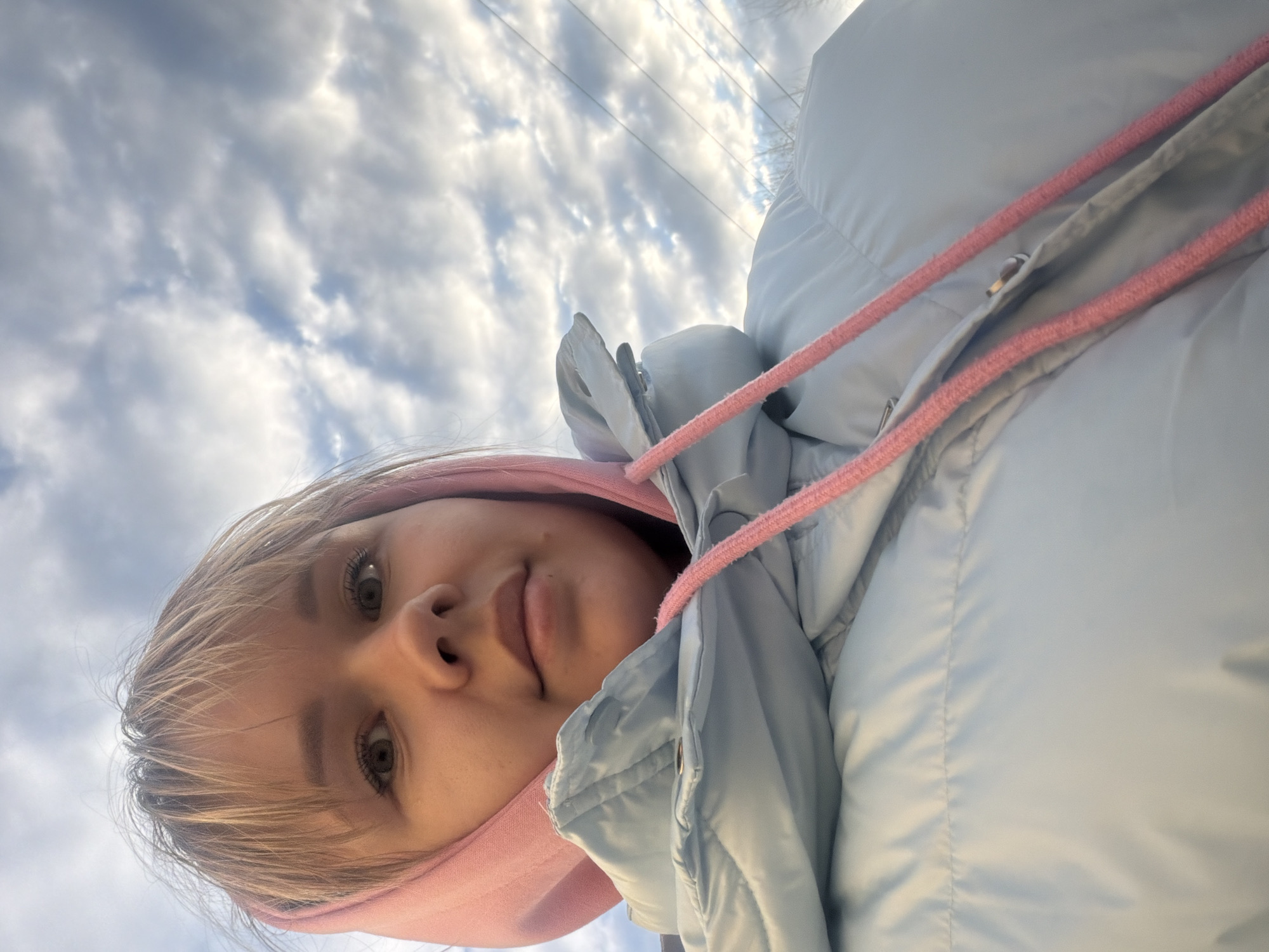 A low-angle, inverted selfie captures a young person, likely female, against a bright, partly cloudy sky. The subject's head and upper torso are prominent, positioned upside down relative to the horizon. She has light-colored hair framing her face, and her eyes are open, looking directly towards the camera. Her expression is neutral and calm. She is wearing a light blue puffer jacket over a pink hooded sweatshirt. A vibrant pink drawstring from the hoodie is visible across the front of her jacket. The background is dominated by a bright blue sky with numerous white cumulus clouds, indicating daytime with fair weather. Faint dark lines, possibly power lines or bare tree branches, are subtly visible in the distance against the sky. The unusual perspective suggests the photo was taken from below, looking up. No text is visible in the image.