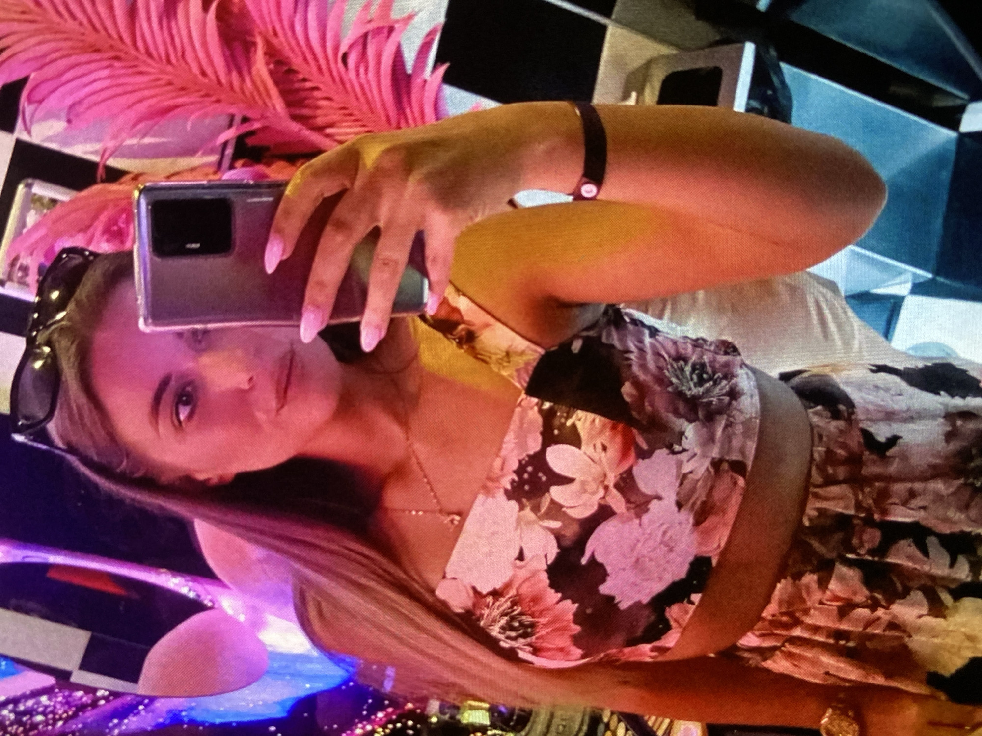 A young woman takes a selfie in an indoor setting, likely an evening venue such as a bar, club, or restaurant in Bankya, Bulgaria. She is positioned on her side, holding a dark-colored smartphone above her head with her right hand to capture the image. Her nails are painted a light pink.
She has long, straight blonde hair and is wearing sunglasses on her head. Her attire includes a floral crop top with shades of pink, black, and white, and a matching skirt or shorts. A delicate necklace with a small pendant is visible around her neck, and she wears a dark bracelet on her left wrist. Her gaze is directed towards the camera with a calm and confident expression.
The background features a prominent black and white checkerboard pattern, possibly on a wall or furniture. Large, decorative pink artificial foliage, resembling feathers or palm leaves, is visible above and behind her head. The scene is illuminated by ambient artificial lighting, casting purple, blue, and pink hues across the environment, suggesting a vibrant, nocturnal atmosphere. No discernible text is visible within the image.