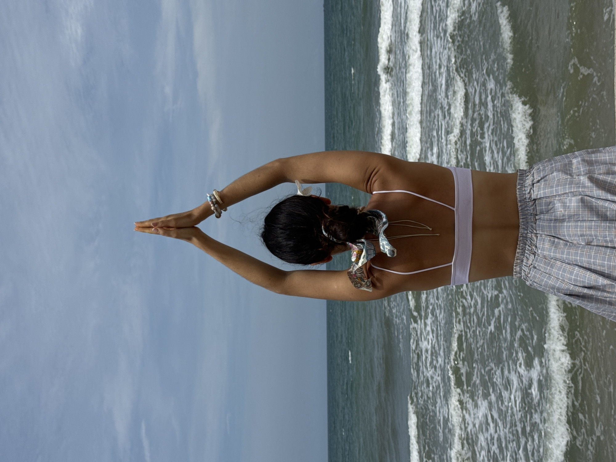 A person is seen from behind, standing on a beach with their arms raised above their head and palms pressed together in a prayer-like gesture. They are wearing a white bralette and a grey and white checkered skirt. The person's hair is pulled back into a bun with a patterned scrunchie. The ocean is visible behind them, with waves rolling towards the shore under a cloudy sky. The scene appears to be located in Phu Thuy Ward, Vietnam.