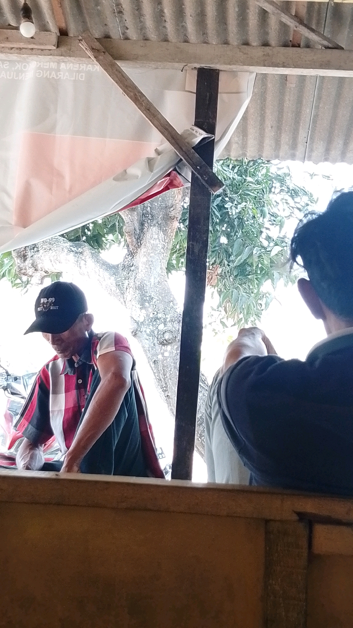 A medium shot captures two individuals outdoors, possibly in Mojopetung, Indonesia. The person on the left, wearing a black cap with "JO-89" printed on it and a red and white checkered shirt, is bending over. Their right arm is extended, and their hand appears to be holding a white object, possibly a bag or cloth. In the background, a tree with textured bark is visible, along with lush green foliage. The sky is bright, suggesting daylight. The other individual, on the right, is seen from behind, wearing a dark blue shirt. They are also looking towards the area where the first person is working. The scene is set under a corrugated metal roof supported by wooden and metal structures. A white banner with red lettering is draped behind the individuals. The overall atmosphere is casual and suggests a working or casual gathering.