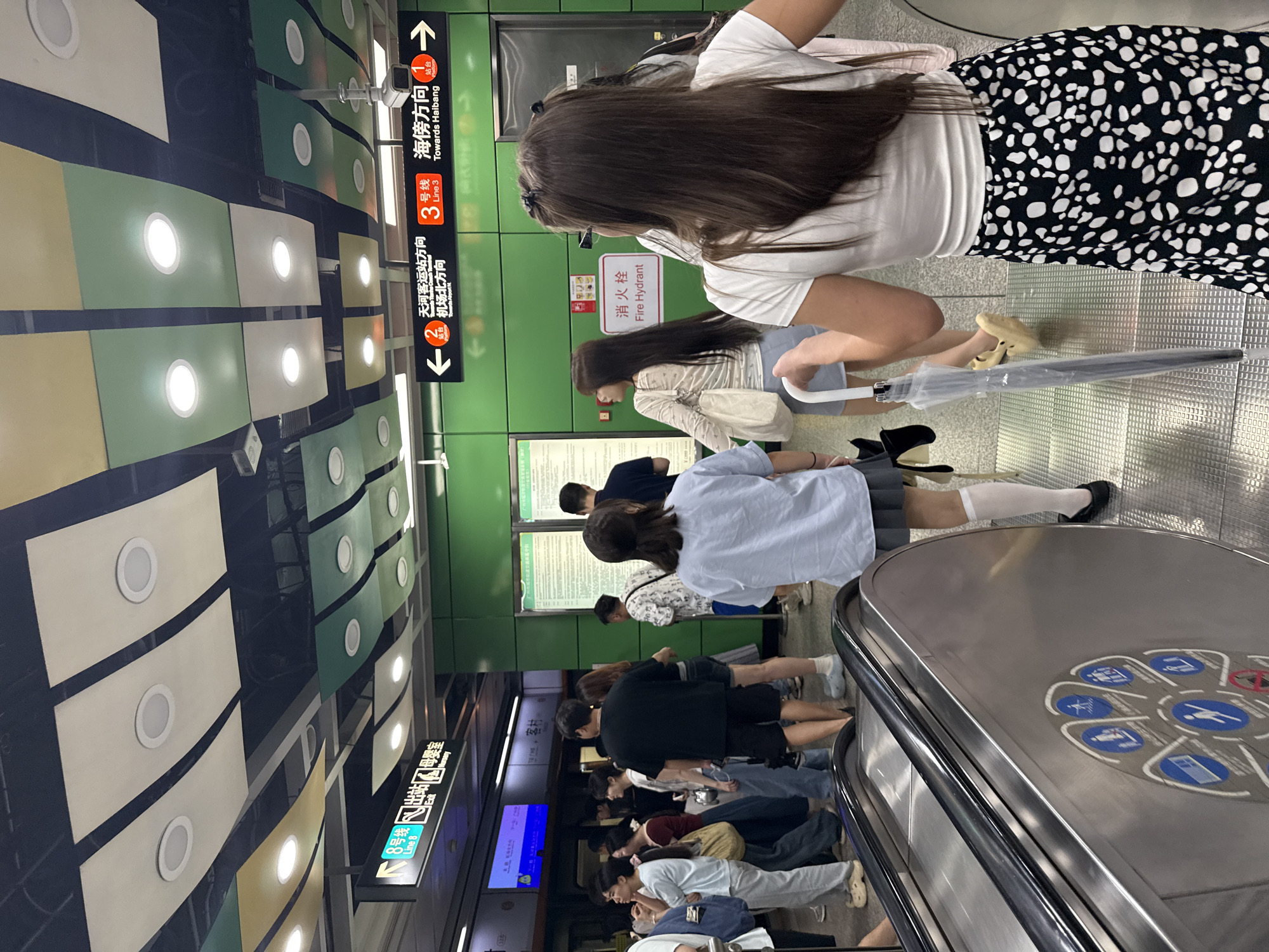 The image depicts an indoor scene, likely a subway station in Haizhu District, China, given the signage. The environment features a green wall with prominent directional signs indicating subway lines and destinations. The ceiling is elaborately decorated with colorful panels and recessed lighting. In the foreground, an escalator is partially visible, with a circular decal featuring various icons, possibly related to station amenities or safety guidelines. Several individuals are present, some standing and others in motion. One woman in the foreground, facing away from the camera, wears a white t-shirt and a black and white patterned skirt. Another woman in the mid-ground, also facing away, is wearing a light blue shirt and a grey skirt, holding a clear umbrella. Other people are scattered throughout the scene, appearing to be commuters. The overall atmosphere suggests a busy public transportation hub.