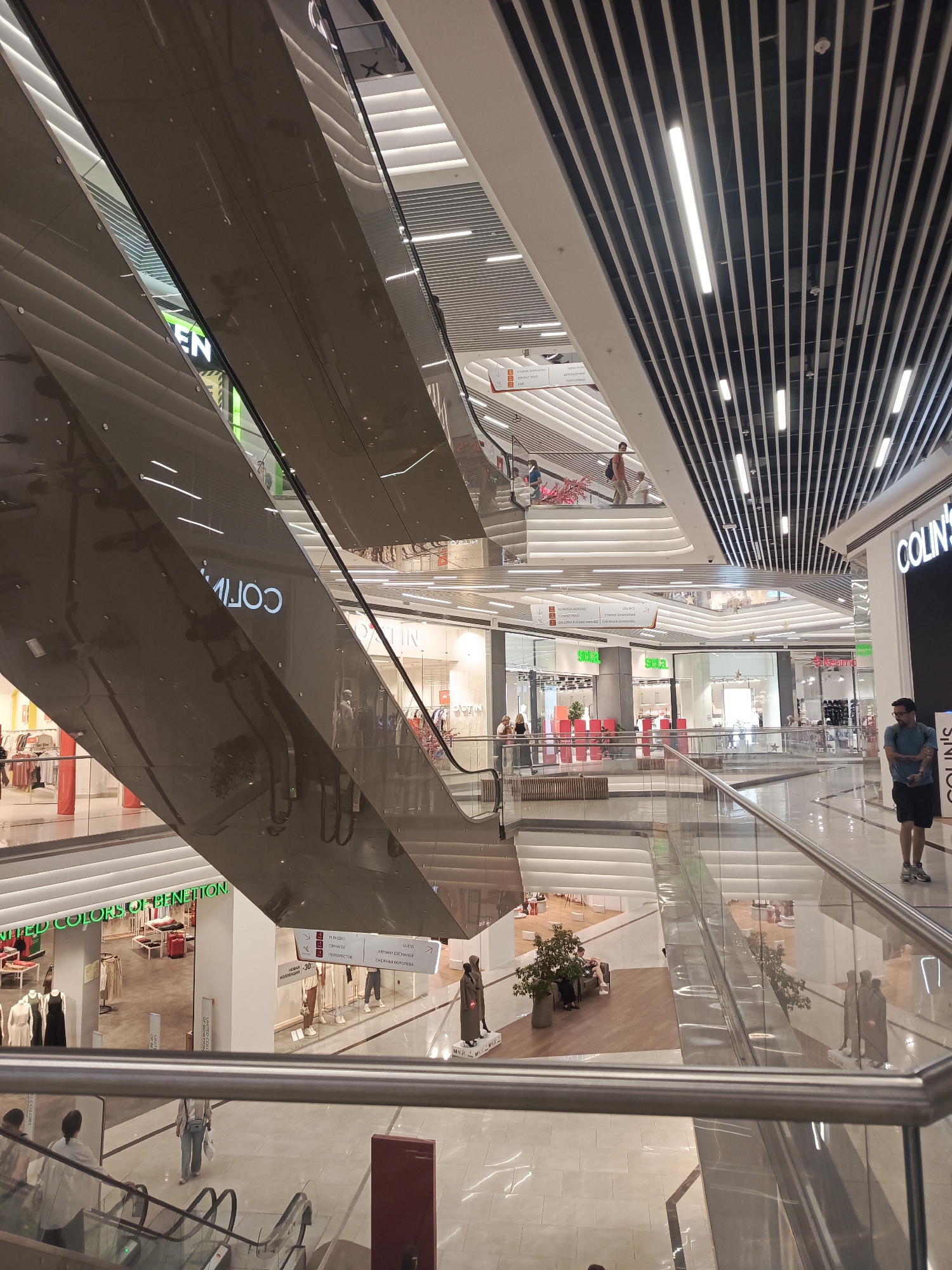 The image depicts the interior of a multi-level shopping mall. Several escalators are visible, with people using them or standing nearby. Stores with visible signage such as "UNITED COLORS OF BENETTON" and "COLIN'S" are present. The architecture features modern design with extensive use of glass, metal, and bright lighting. The flooring appears to be light-colored tile. The scene is well-lit, suggesting daytime or well-illuminated interior spaces. The environment is clean and organized, characteristic of a commercial shopping center. The photograph was taken inside the "Europe" shopping center in Perm, Russia.