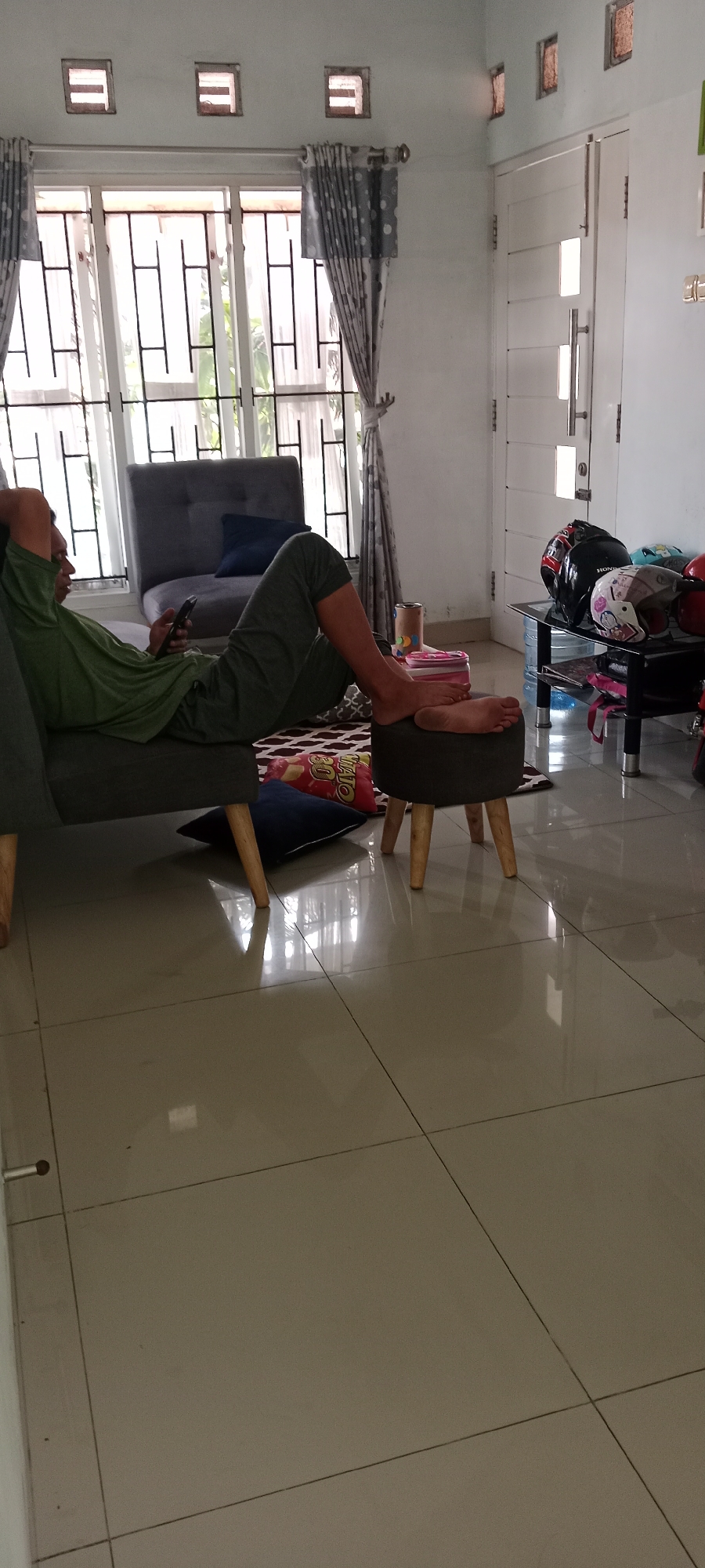 The image depicts an indoor scene, likely a living room, within a residence in Maguwoharjo, Indonesia. A person is reclining on a sofa, dressed in a green long-sleeved shirt and dark pants, holding a mobile phone. Their legs are propped up on a small ottoman. The room features a large window with patterned curtains and metal bars on the exterior, suggesting security measures. A dark gray armchair is positioned near the window. To the right of the frame, a glass-top table holds several helmets, including two on display and one partially visible. A white door with vertical paneling is also visible on the right side of the image. The floor is tiled with a beige, glossy finish, reflecting light. Several pillows are scattered on the floor, and a patterned rug is partially visible beneath the ottoman. The lighting suggests an indoor environment during the daytime.