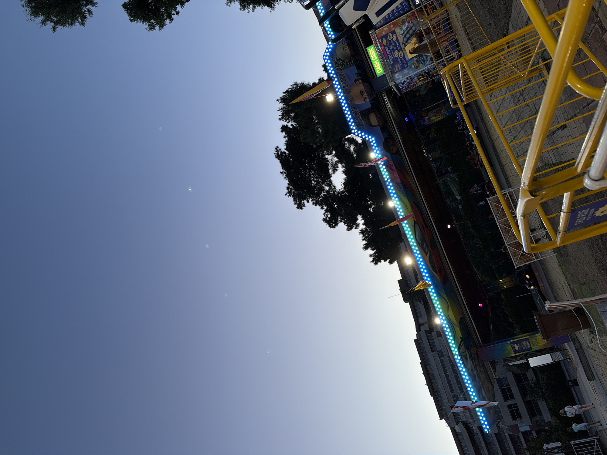The image depicts an outdoor amusement park attraction at dusk. The attraction features a ride with colorful LED lights and graphics of cars and faces. A "CASH DESK" sign is visible on the attraction, along with pricing for tokens. In the foreground, a yellow metal barrier encloses a pathway. In the background, a large, multi-story building is visible, along with trees. Two young females are walking on the pathway near the building. The sky is a clear, pale blue, indicating either early morning or late evening.