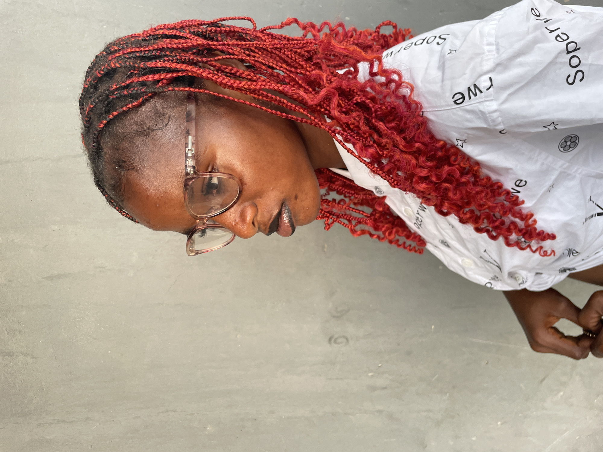 A close-up, side-profile portrait captures a person, likely female, with dark skin and vibrant red hair. The hair is styled in thin braids at the roots, transitioning into voluminous, curly red extensions that cascade over the shoulders. The person is wearing clear-framed glasses with a subtle pinkish tint. A dark, possibly metallic, lip color is visible on their lips.
They are dressed in a white collared shirt, which features repeating black text and patterns. The visible text appears to be "Soperwe" or "Sopwerwe," along with small star symbols and a circular emblem that looks like a patterned sphere or flower. The shirt has folds and creases, suggesting movement or relaxed wear.
The person's gaze is directed downwards, creating a contemplative or focused expression. In the lower right corner, a hand is partially visible, appearing to be gently clasped or holding something, adorned with simple rings.
The background is a plain, light grey surface, possibly a wall or concrete floor, providing a neutral backdrop that highlights the subject. The lighting is even and bright, suggesting daytime conditions. The overall scene conveys a calm and observant mood, captured in Badagry, Nigeria.