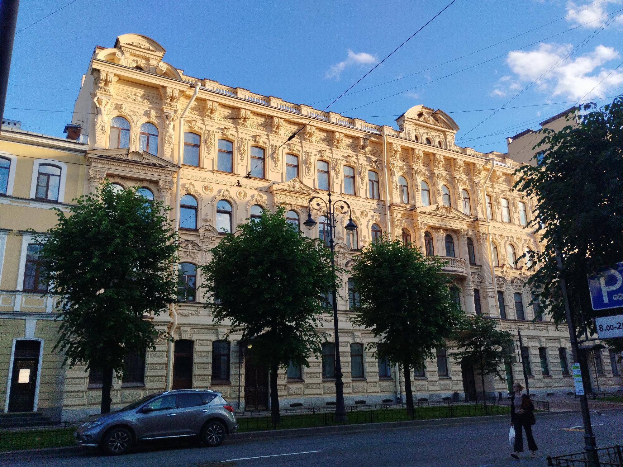 The image shows a wide shot of a cream-colored building with ornate architectural details, including sculptures, arches, and decorative friezes, located in Saint Petersburg, Russia. The building has multiple stories and numerous windows, many of which are arched. Green trees line the street in front of the building, with a few trees partially obscuring the facade. A grey SUV is parked on the side of the street. A woman in dark clothing is walking across the street, carrying a white plastic bag. A parking sign with the letter "P" and time restrictions is visible on a pole near the right edge of the image. The sky is clear blue with a few wispy clouds, suggesting it is daytime. The overall scene depicts a street view in a historic urban environment.