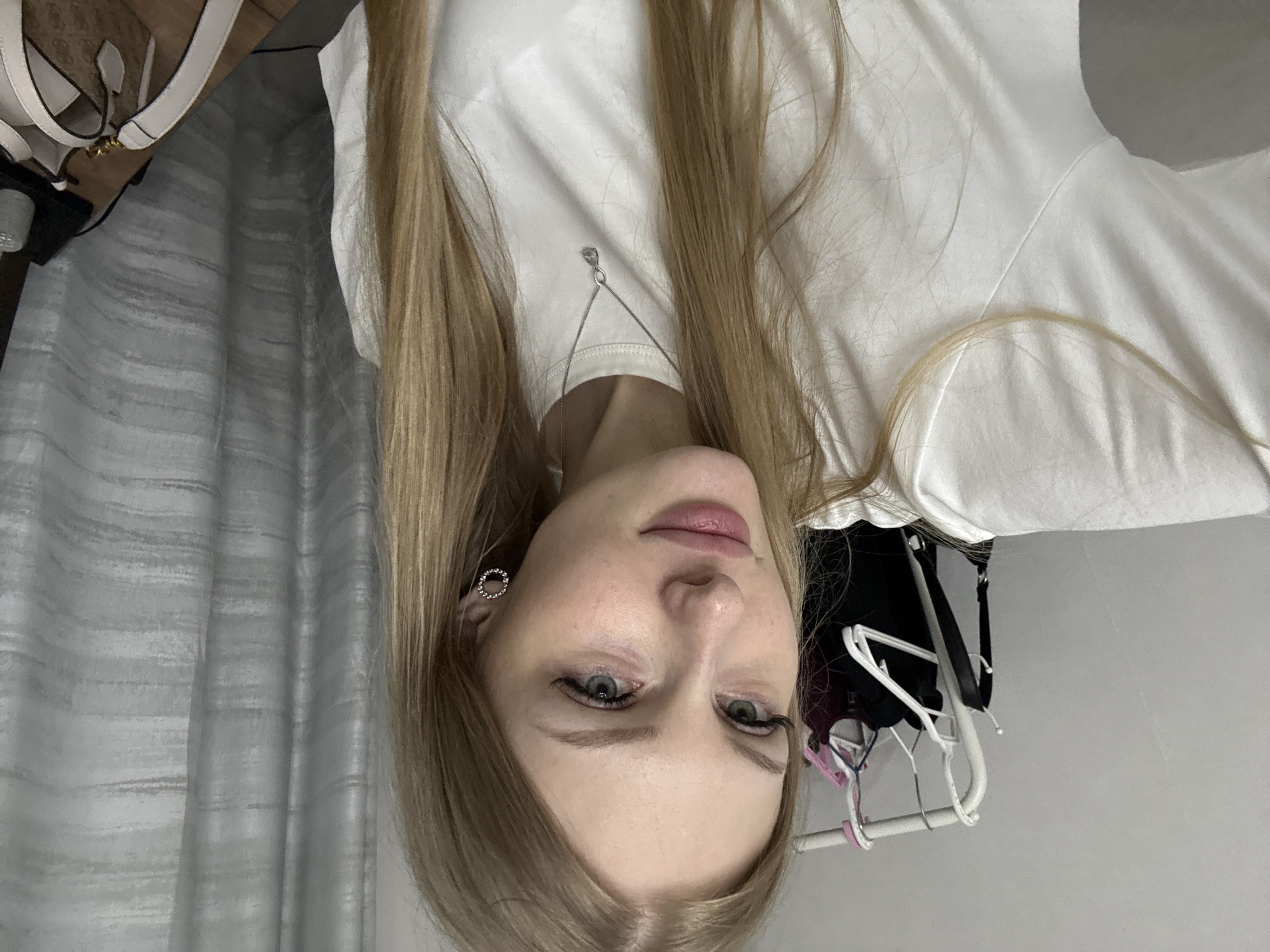 A young Caucasian woman with blonde hair and blue eyes is posing for a selfie in what appears to be a bedroom. She is wearing a white t-shirt and a silver necklace with a teardrop pendant. She has on circular, silver earrings. In the background, a clothing rack with several garments is visible. The room also features a grey curtain and a wooden surface with a bag on it. The overall impression is casual and personal. The image does not provide enough information to determine the specific location within the city of Perm, Russia.