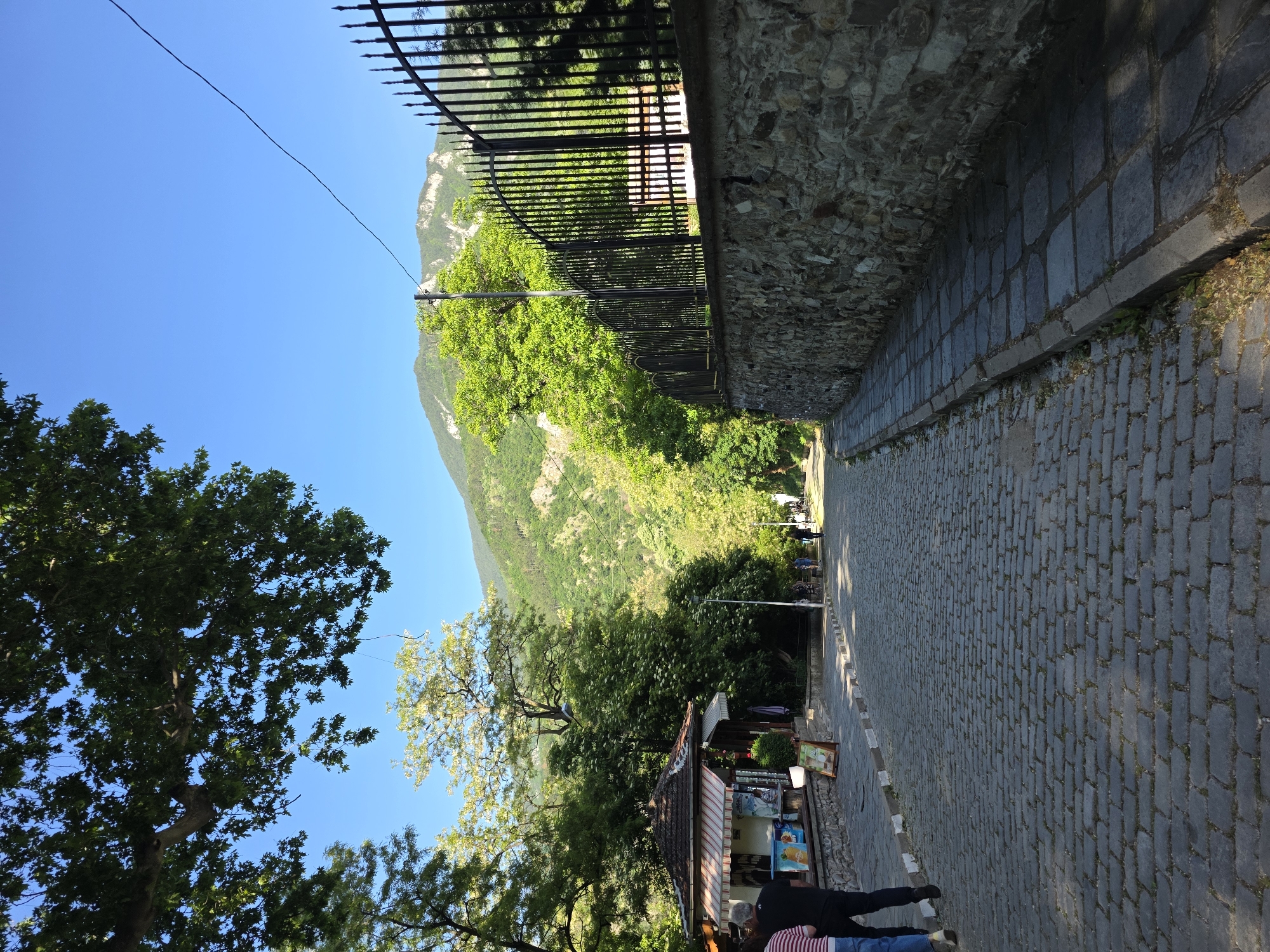 This picture shows a street in the city of Bachkovo, Bulgaria, lined with shops and vegetation. The street itself is made of cobblestone and ascends slightly. Along the right of the street is a stone wall with a metal fence on top. On the left, there are trees and a small shop. Two people are visible in the foreground. In the distance, a mountain is visible under a clear sky.