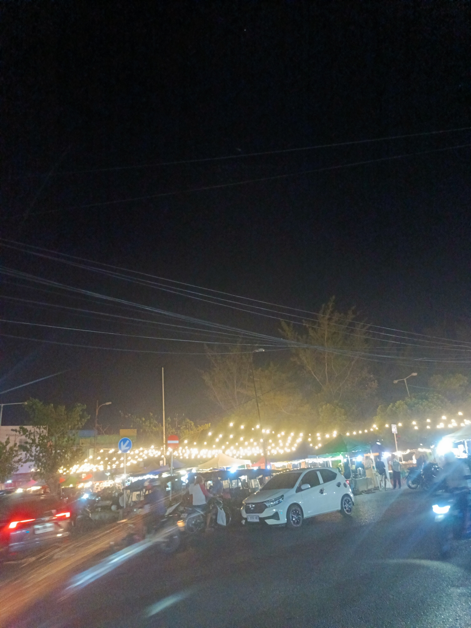 This is a night-time photograph of a street market or gathering in Luwuk, Indonesia. The scene is illuminated by numerous string lights draped across the area, creating a warm, festive ambiance. In the foreground, a white car is parked, and to its left, a car with its red taillights illuminated is moving. Several motorcycles are also present, with one being ridden and another parked. People are visible, some standing and interacting, while others appear to be walking or attending to stalls. Tents or market stalls are set up, indicating a commercial or community event. The background features trees and streetlights, along with overhead power lines that are prominent against the dark night sky. The overall impression is of a lively evening scene in a public space within the city.