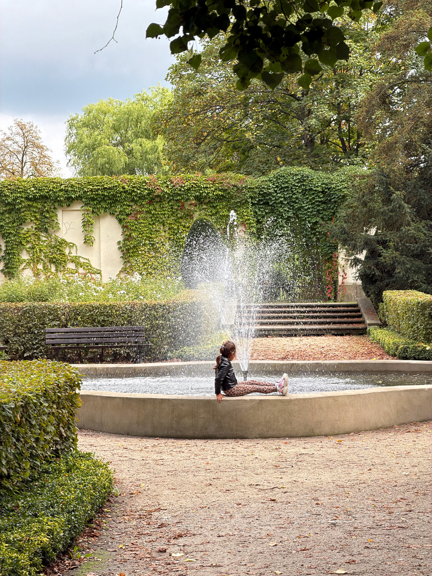 In Wrocław, Poland, a young girl sits at the edge of a circular fountain, her legs extended towards the water. She is wearing a black leather jacket and patterned leggings. The fountain's spray arcs in front of her. Behind the fountain, a wall is covered in lush green ivy, with some red foliage interspersed. Steps ascend to a higher level behind the wall. To the left of the fountain, a dark wooden bench is visible amidst manicured bushes. The ground surrounding the fountain is a gravel path scattered with fallen leaves. The scene is set in a park-like environment with mature trees in the background.