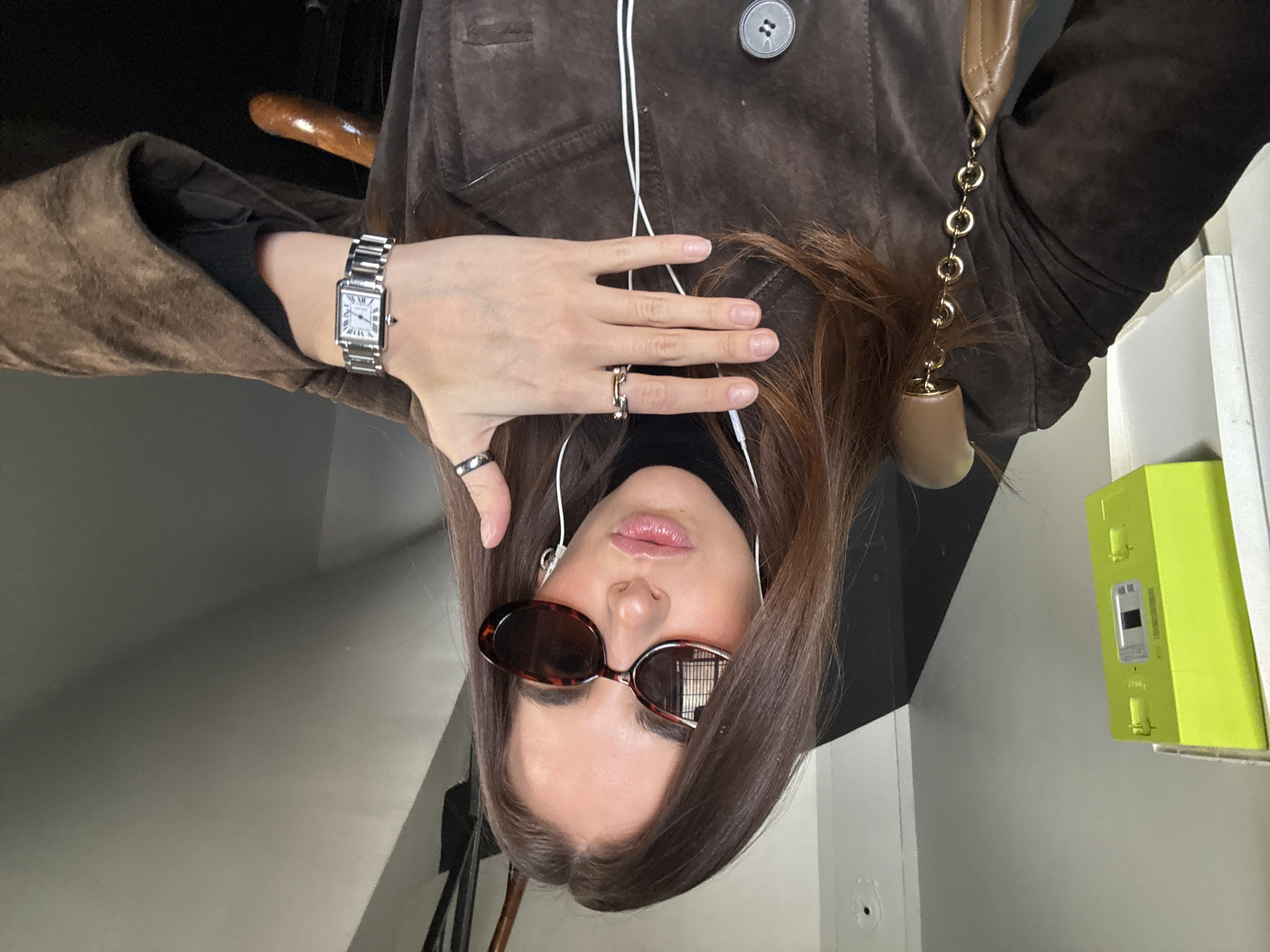 A female individual, approximately 20-35 years old, is depicted in a selfie, posing indoors in what appears to be a stairwell or hallway in Paris, France. She has long brown hair, wears oval-shaped tortoise-shell sunglasses, and has light pink lipstick applied. White wired in-ear headphones are visible. She is dressed in a dark brown, suede-like jacket over a black turtleneck garment. Her left hand is raised towards the camera, palm slightly open, revealing a silver-toned rectangular watch on her wrist and two rings: a two-tone ring on her middle finger and a silver band on her right-hand ring finger (partially visible). A brown leather bag with a gold chain strap rests on her right shoulder. The background features a light grey wall with a bright lime-green utility meter, consistent with a French Linky meter, mounted on the left. A dark wooden handrail is visible on the lower right, suggesting a staircase.