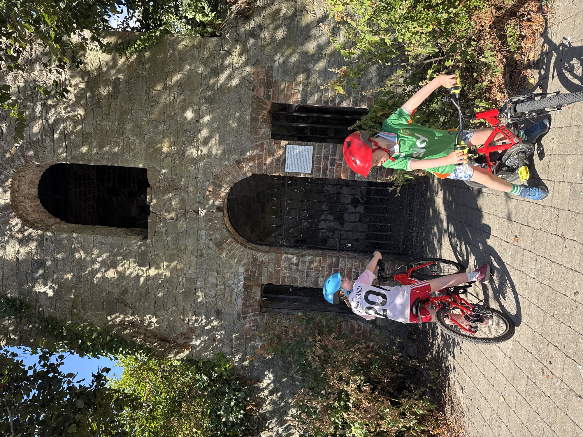Two children, a girl and a boy, are standing with their bicycles in front of an old stone structure in Cahir, Ireland. The girl, on the left, is wearing a light pink t-shirt with the number 20 on the back and red shorts. She has a blue helmet on. The boy, on the right, is wearing a green t-shirt with white trim, patterned shorts, and a red helmet. Both children are facing towards the right side of the frame. The stone structure appears to be an old ruin with arched doorways and windows covered by metal gates. Vegetation, including ivy and bushes, is growing on and around the structure. The ground is paved with concrete slabs. Sunlight casts shadows from the trees and structure onto the scene. A sign is affixed to the wall to the right of the arched entrance. This structure is identified as Cahir's Dayroom, c. 1840.