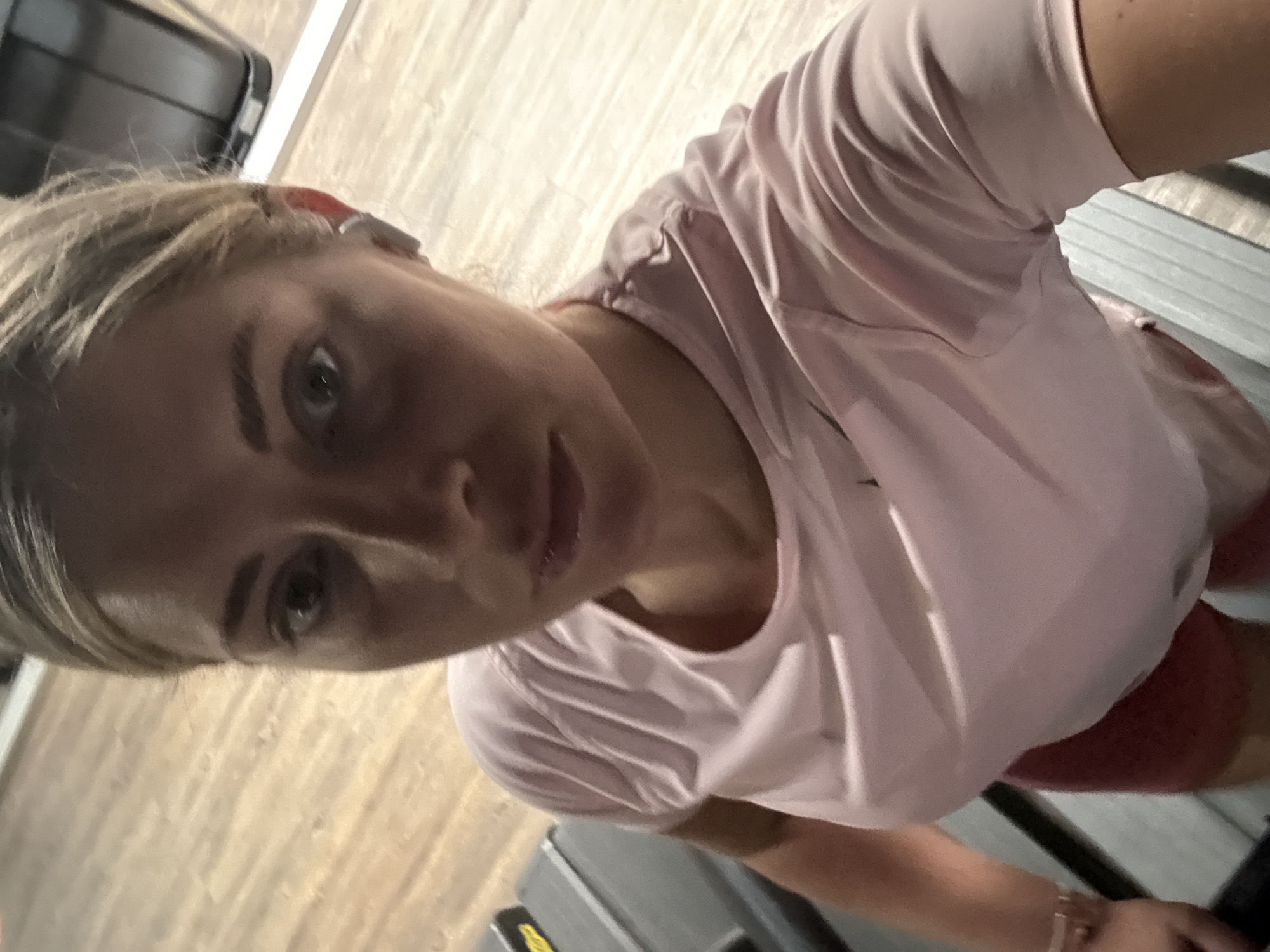 A woman with blonde hair and blue eyes is taking a selfie. She is wearing a light pink t-shirt and red shorts. She appears to be standing on a treadmill. The background is blurred, but the floor appears to be wooden. There is no indication of location.
